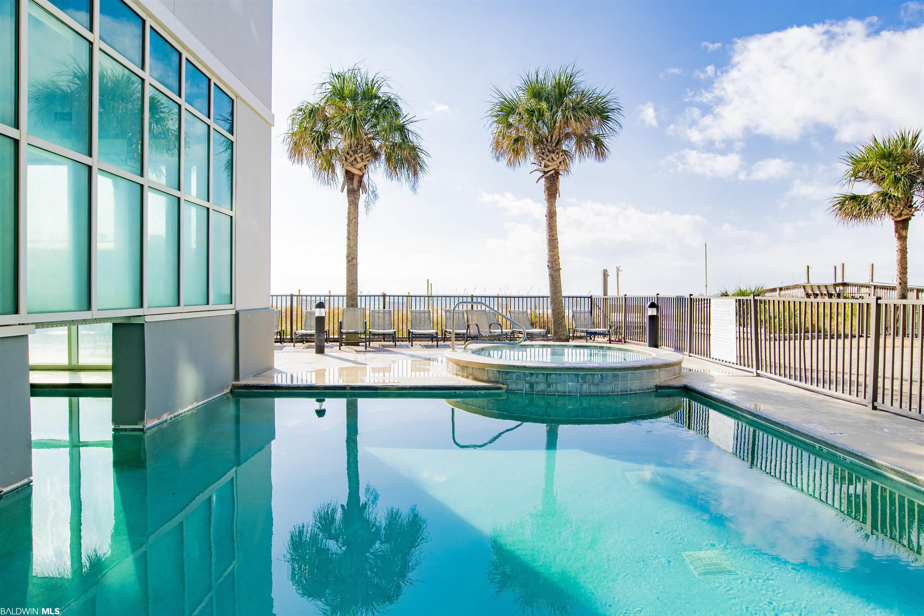Condominium pool and raised spa beside a glass facade, with palm trees and a glimpse of the Gulf.
