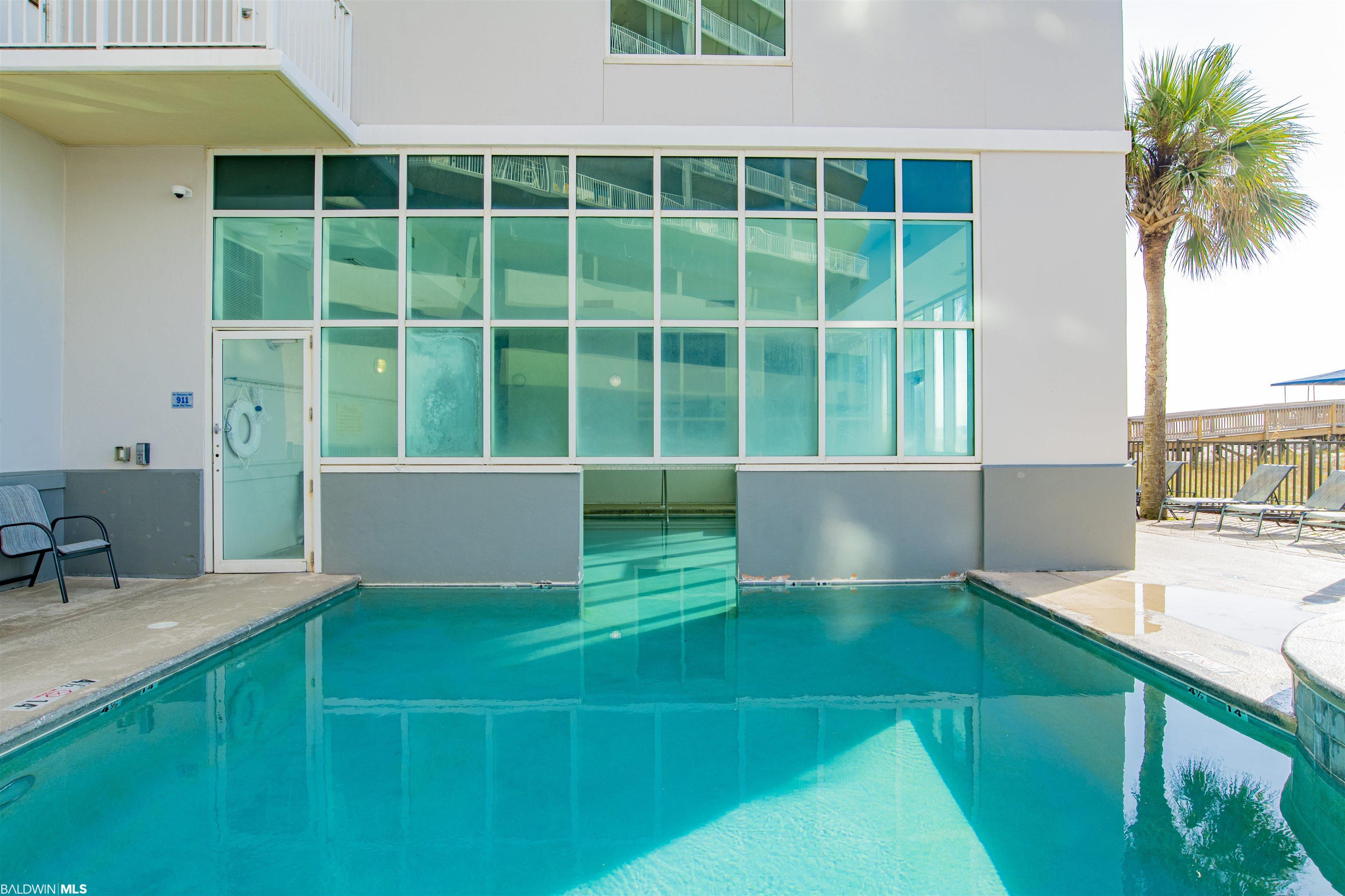Condominium pool beside a glass facade, with a palm tree and poolside lounge chairs.