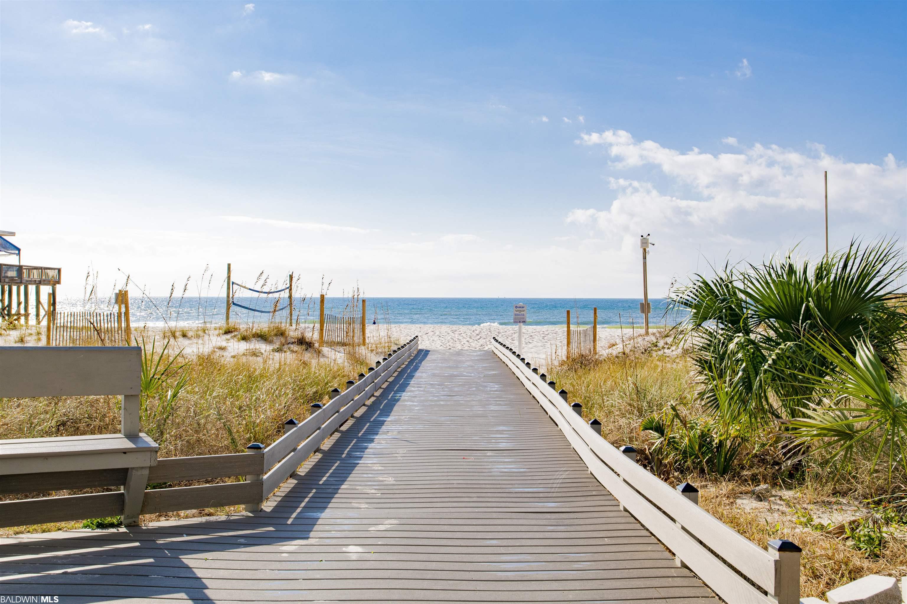 Beach access boardwalk crosses dune grass toward the sandy shore and blue Gulf water.
