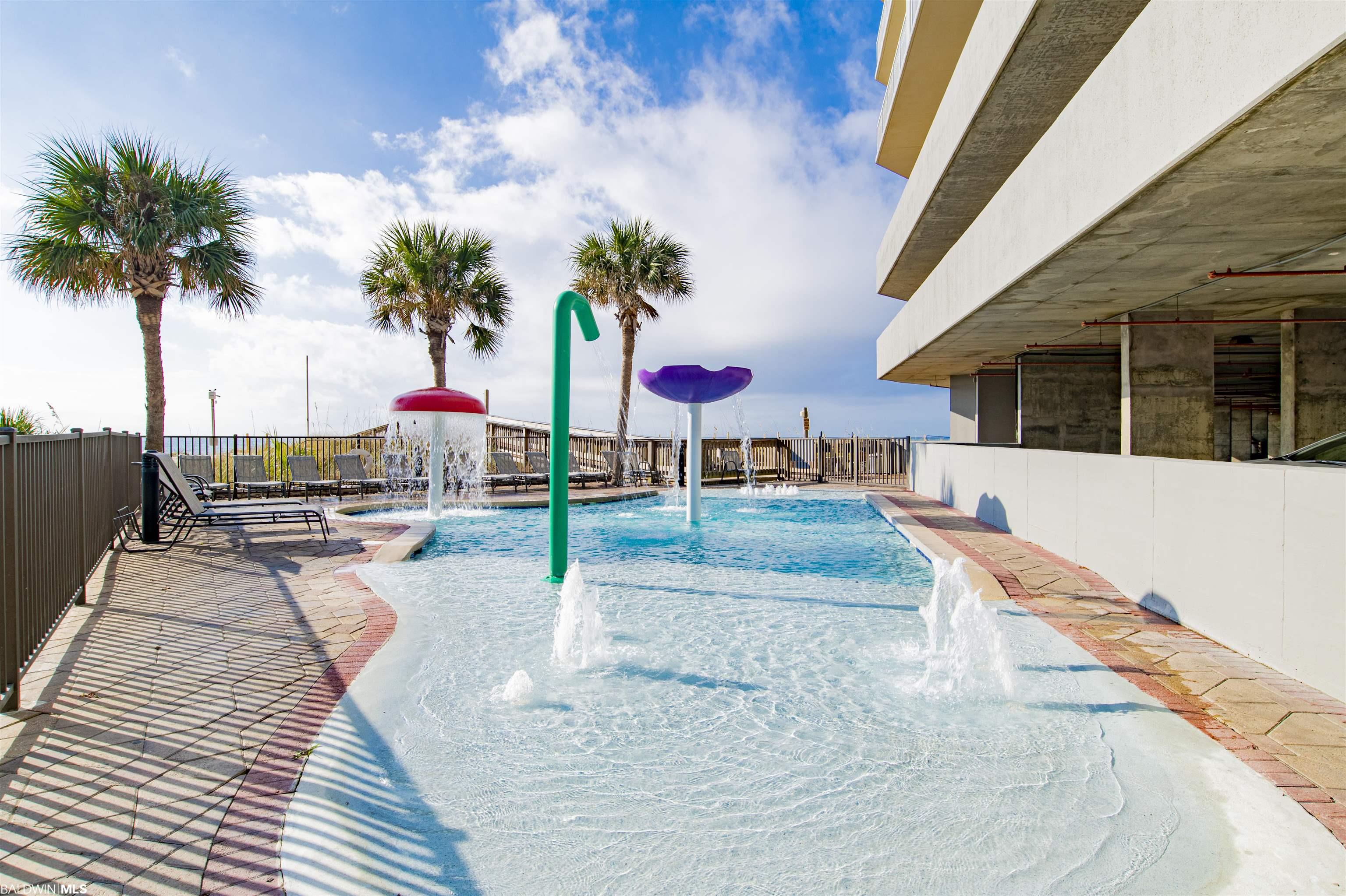 Condominium pool with colorful spray fountains, palm trees, and lounge chairs beside the building.
