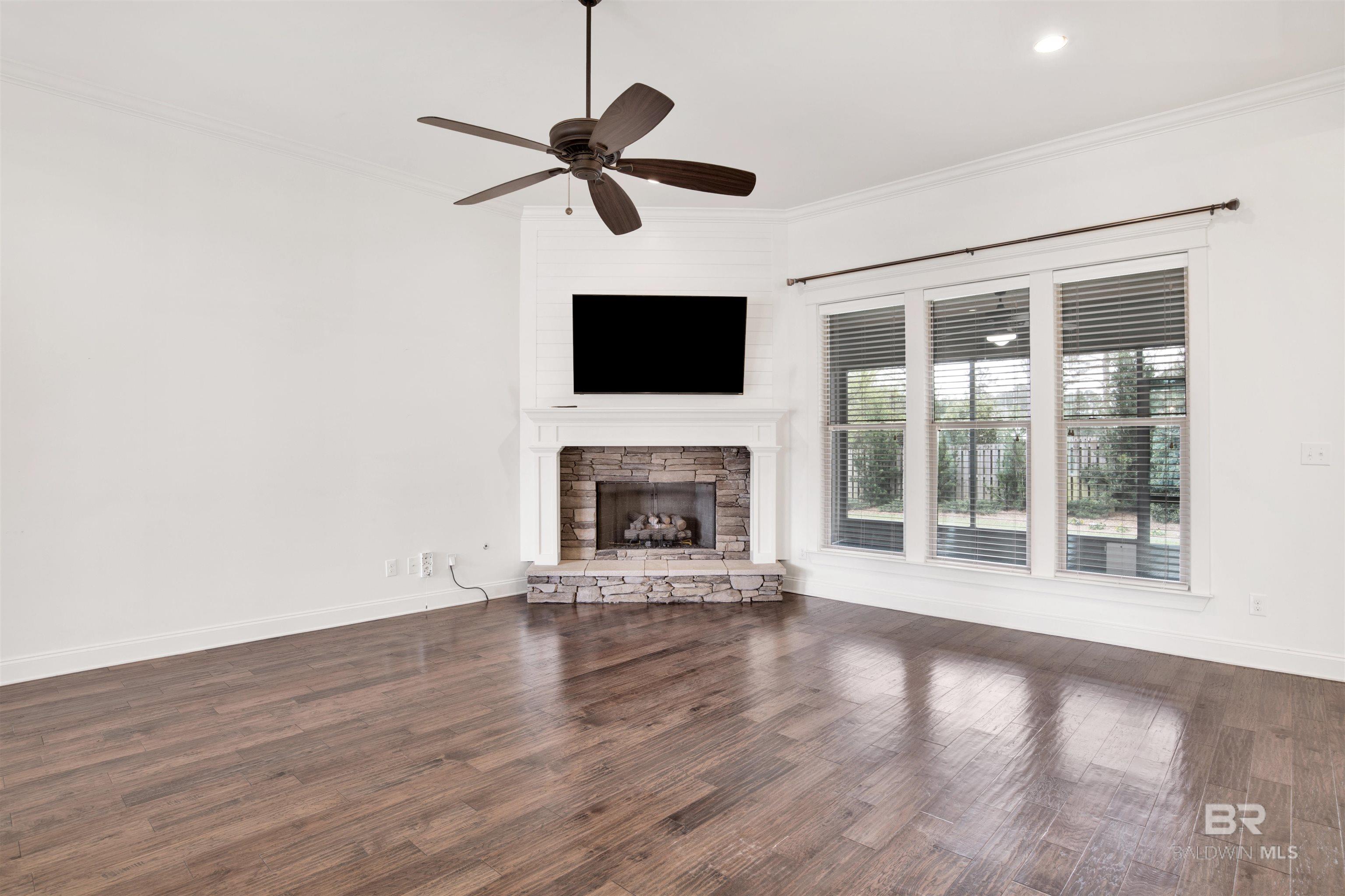 Empty living room with a stone fireplace, wall-mounted television, and large windows.