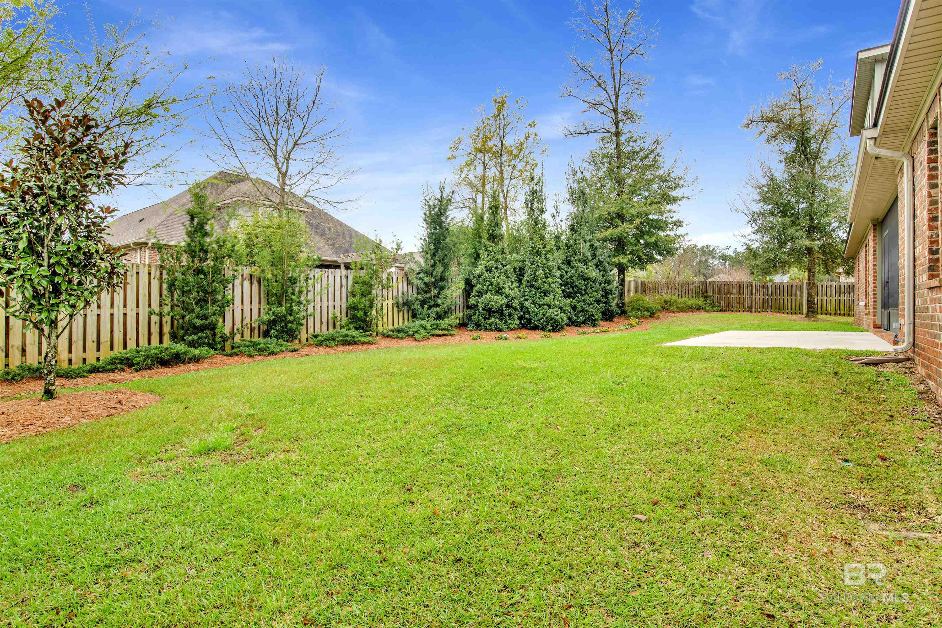 Fenced backyard with a wide lawn, landscaped shrubs, and a concrete patio beside a brick house.