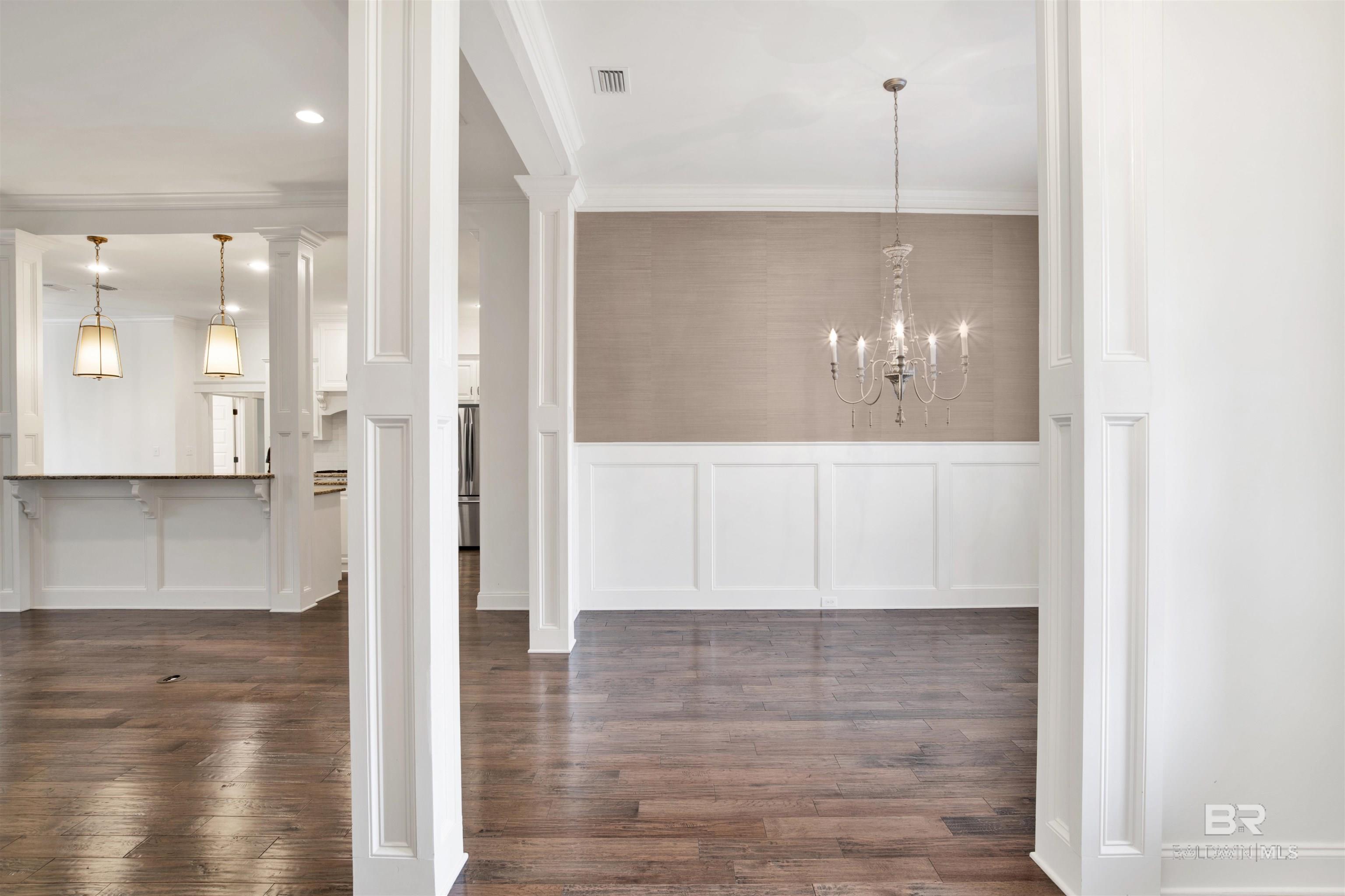 Empty dining room with a chandelier, paneled walls, dark wood floors, and an adjoining kitchen.