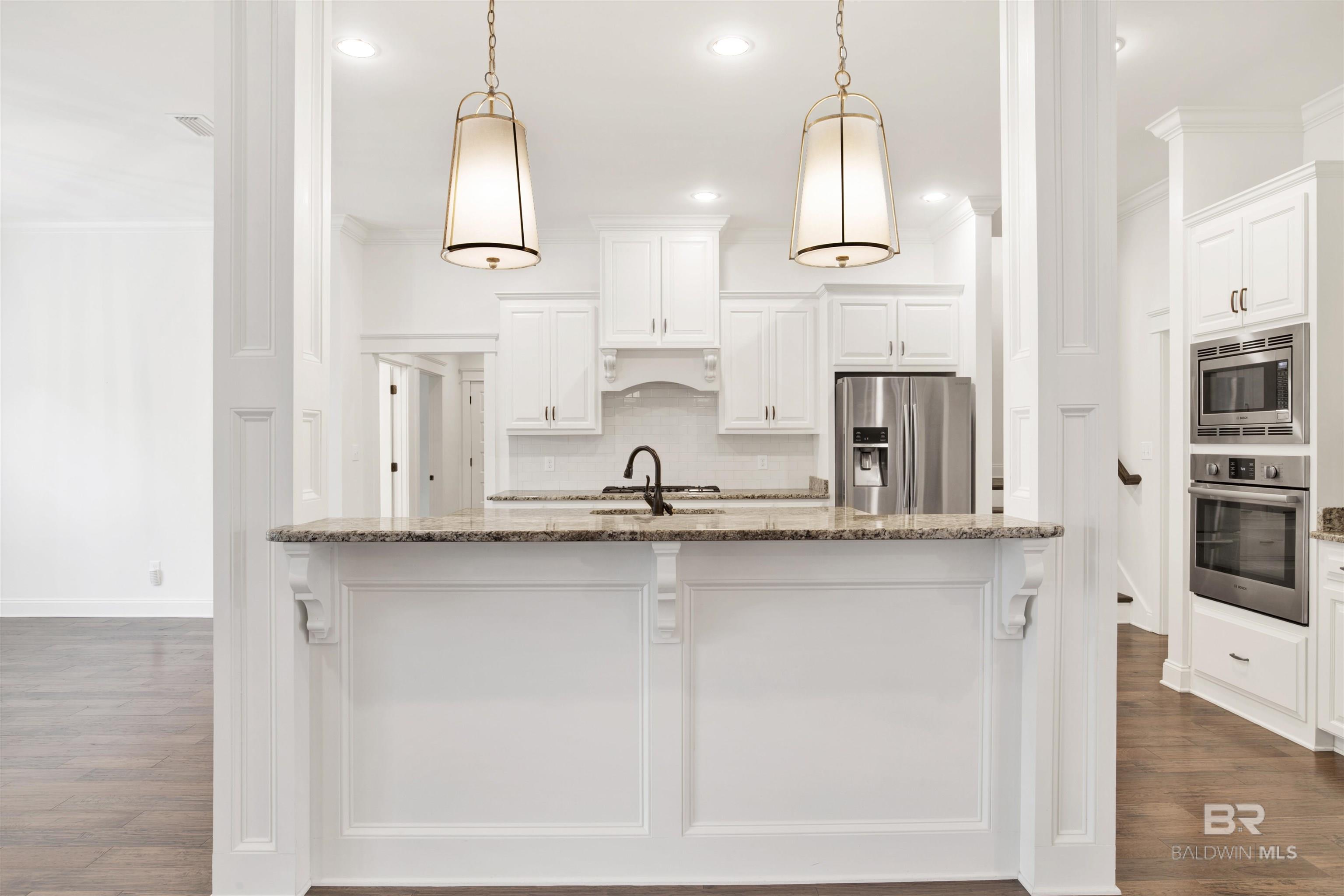 White kitchen with a large granite-topped island, pendant lights, and stainless steel appliances.