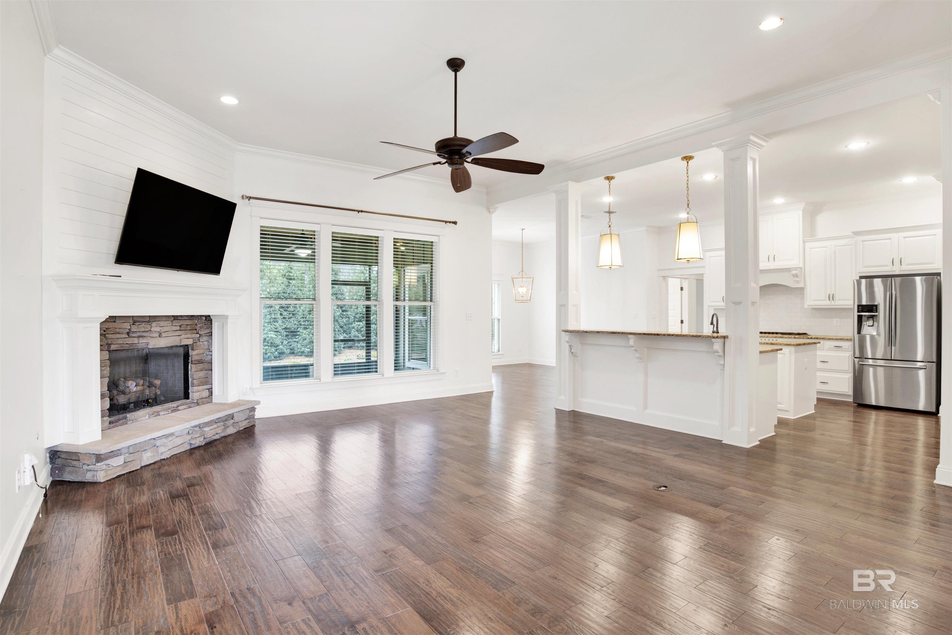 Unfurnished living room opening to a kitchen, with a stone fireplace, large windows, and dark wood floors.