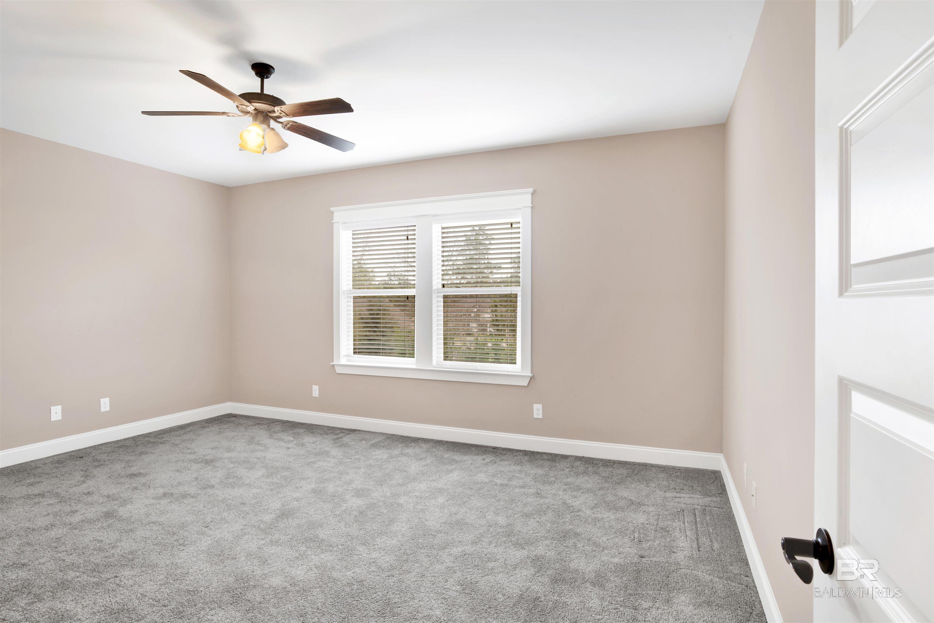 Empty bedroom with gray carpet, beige walls, and a ceiling fan above a window with blinds.