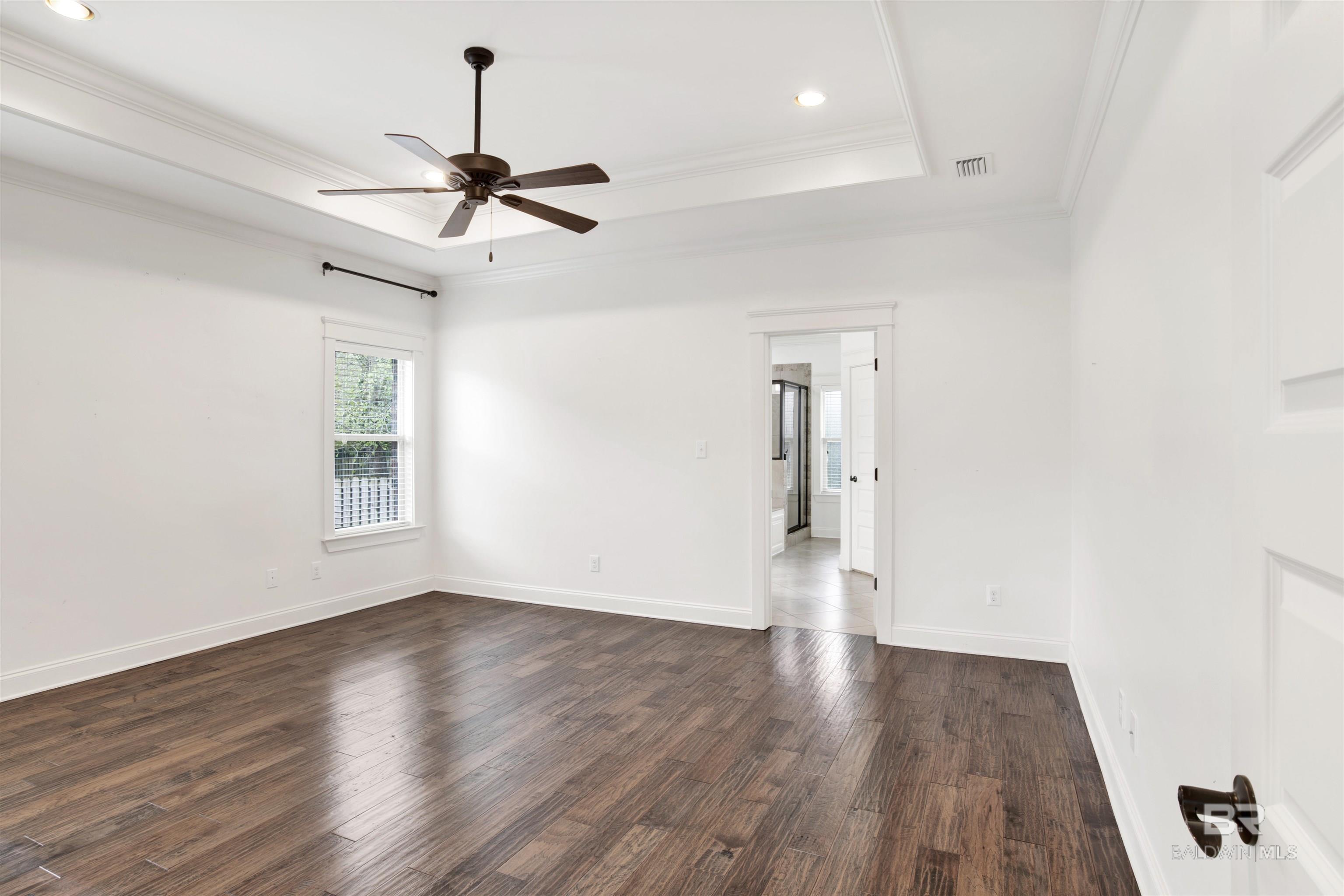 Empty bedroom with dark wood floors, a tray ceiling, and an open doorway to a tiled bathroom.