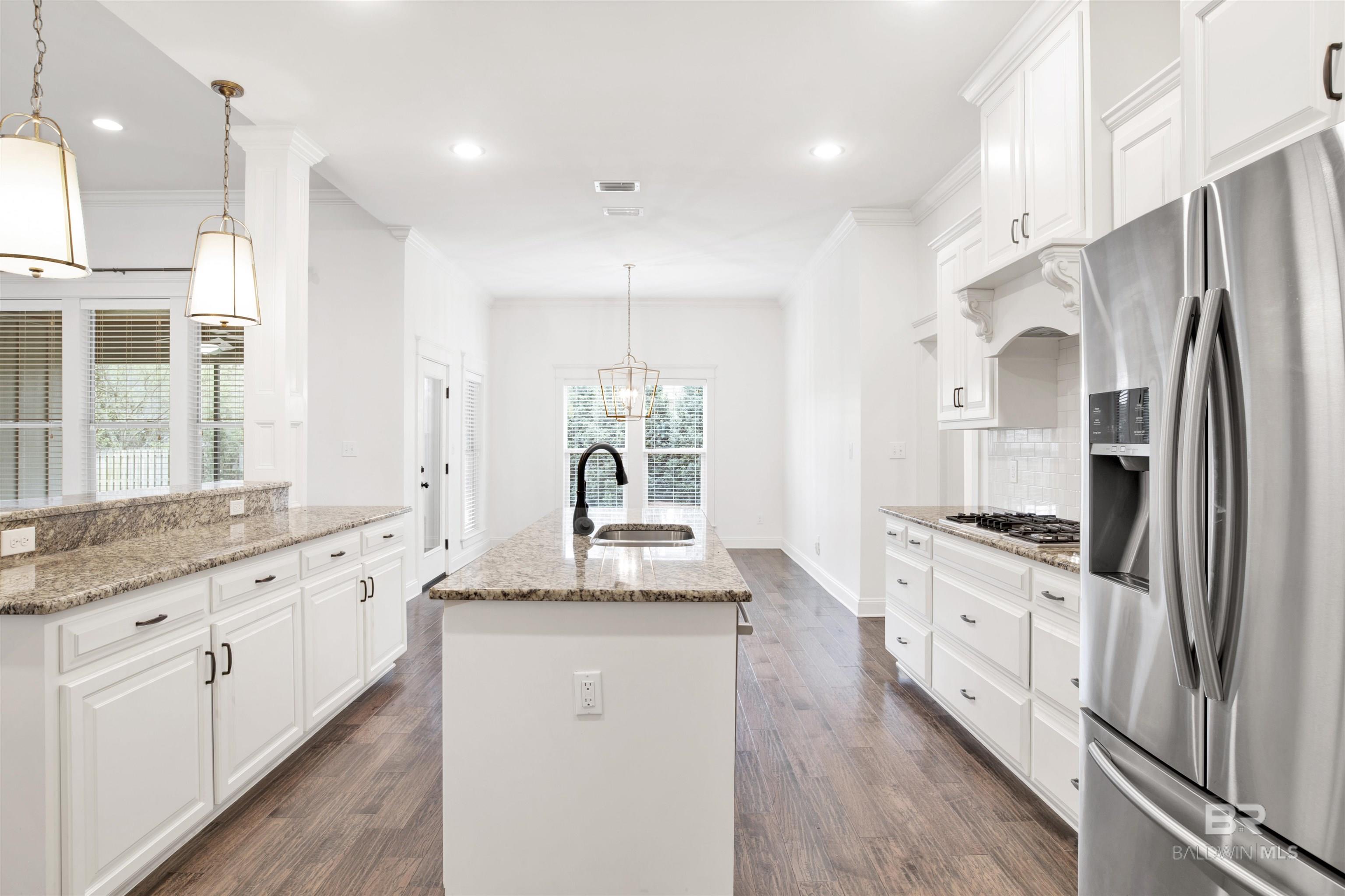 Bright white kitchen with a granite island, gas cooktop, stainless steel refrigerator, and dark wood-look floors.