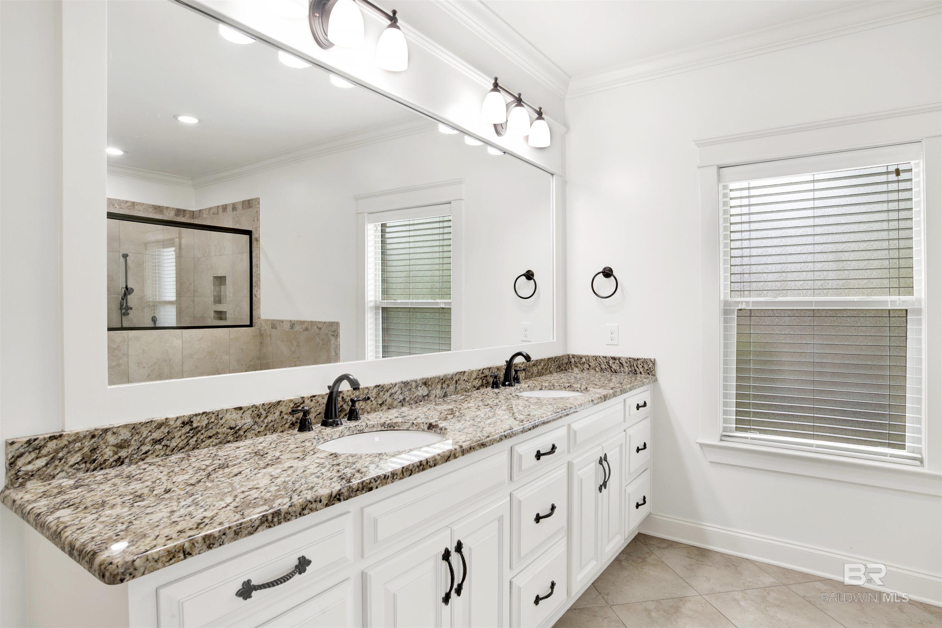 Bathroom double vanity with granite countertops, white cabinetry, and a tiled shower reflected in the mirror.