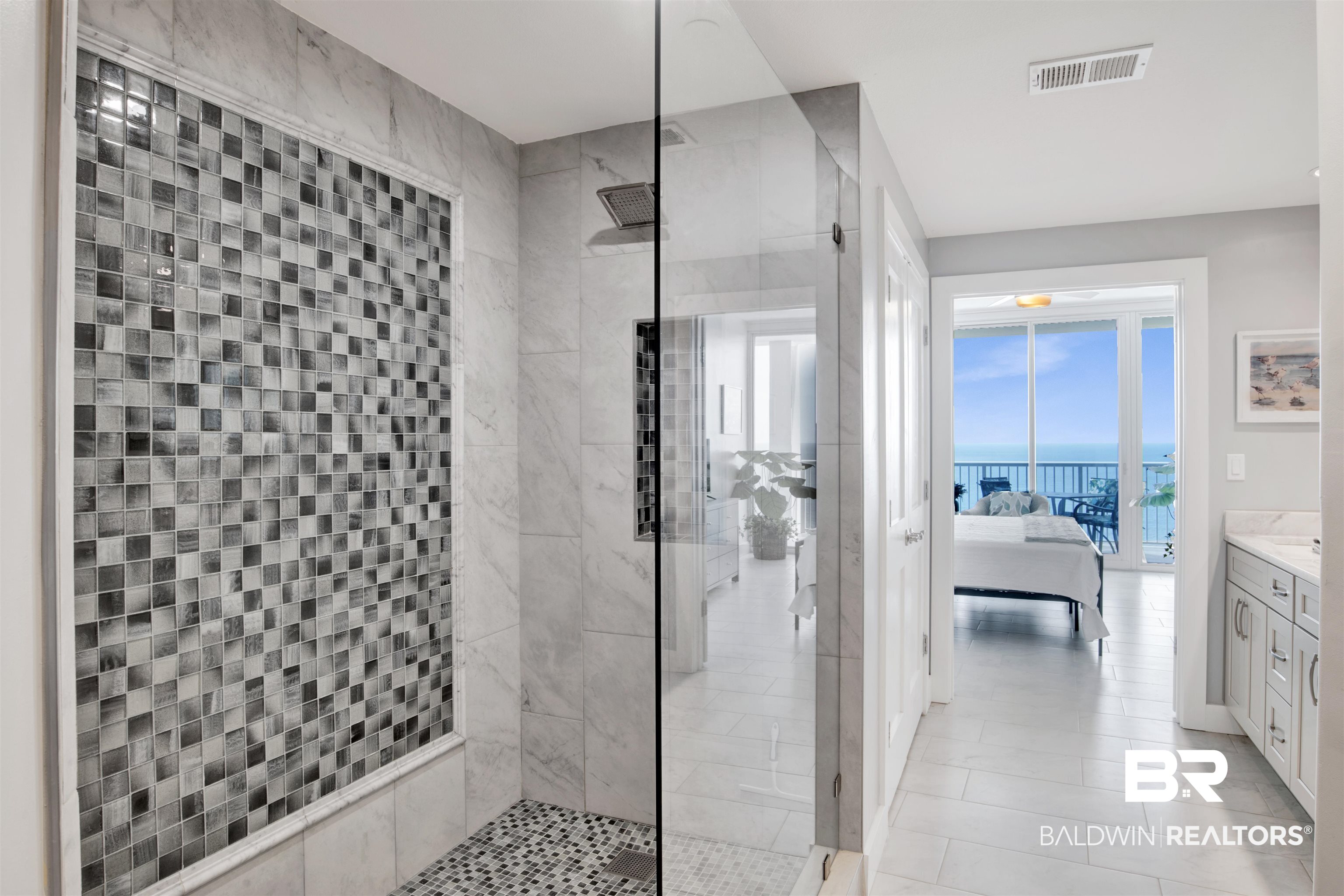 Condo bathroom with a glass shower and mosaic tile wall, opening to an ocean-view bedroom.