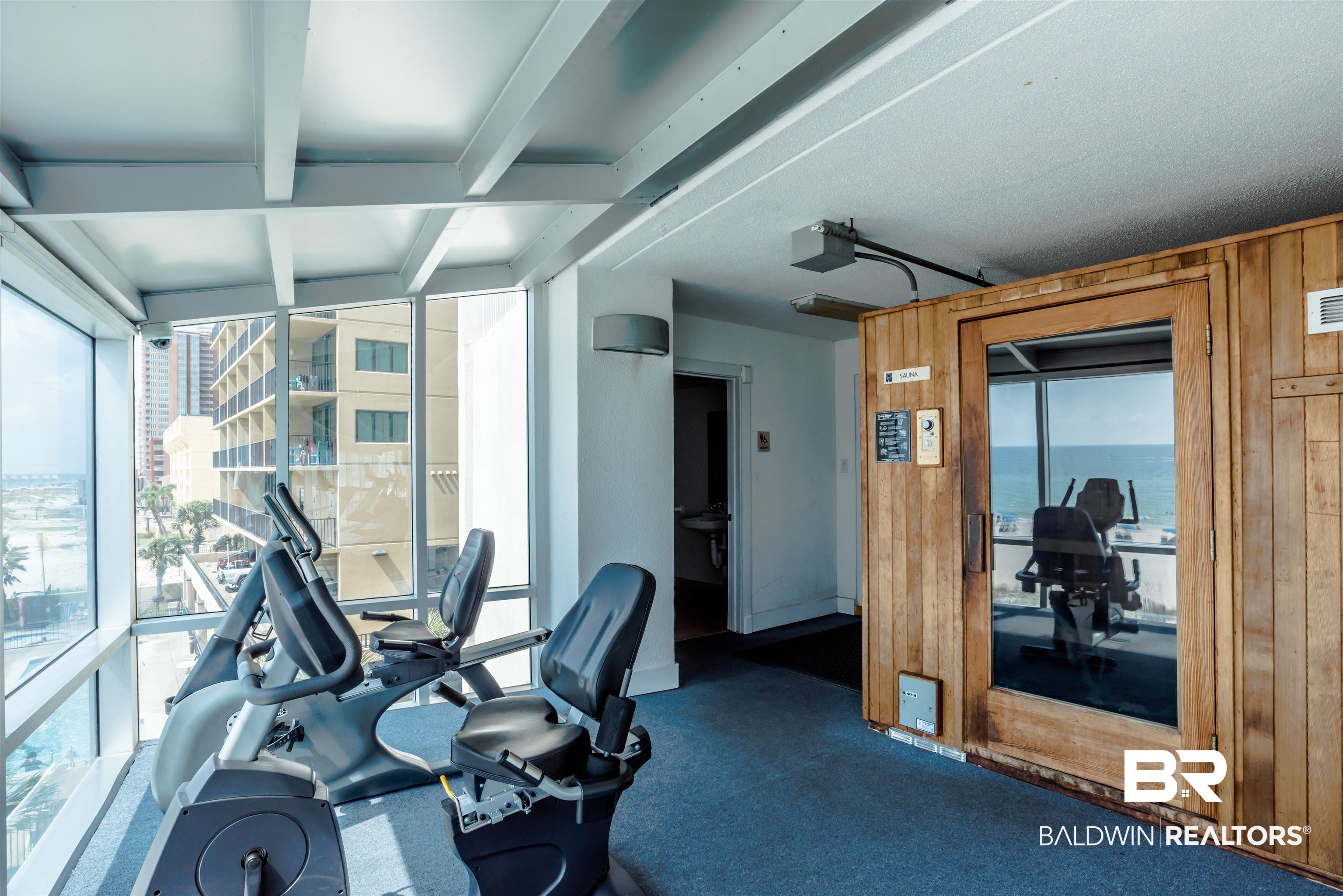 Condo fitness room with exercise machines, Gulf views through tall windows, and a wood-paneled sauna.