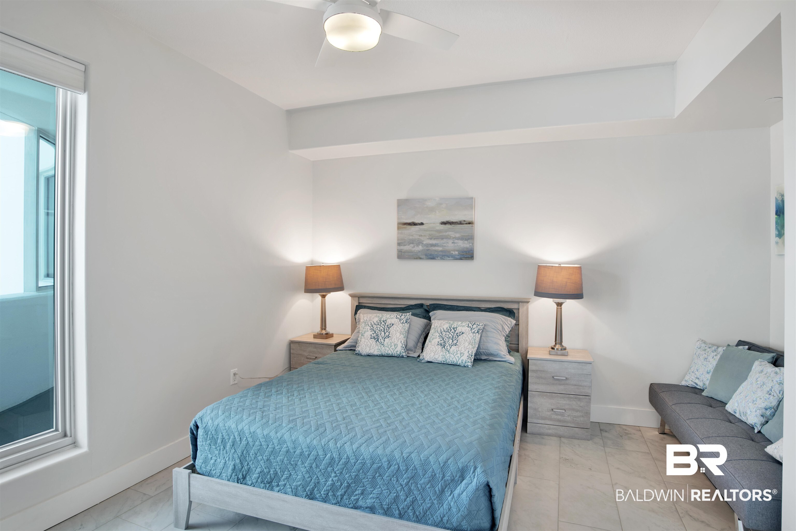 Bedroom with a teal quilted bed, matching lamps, and a gray sofa beside a large window.