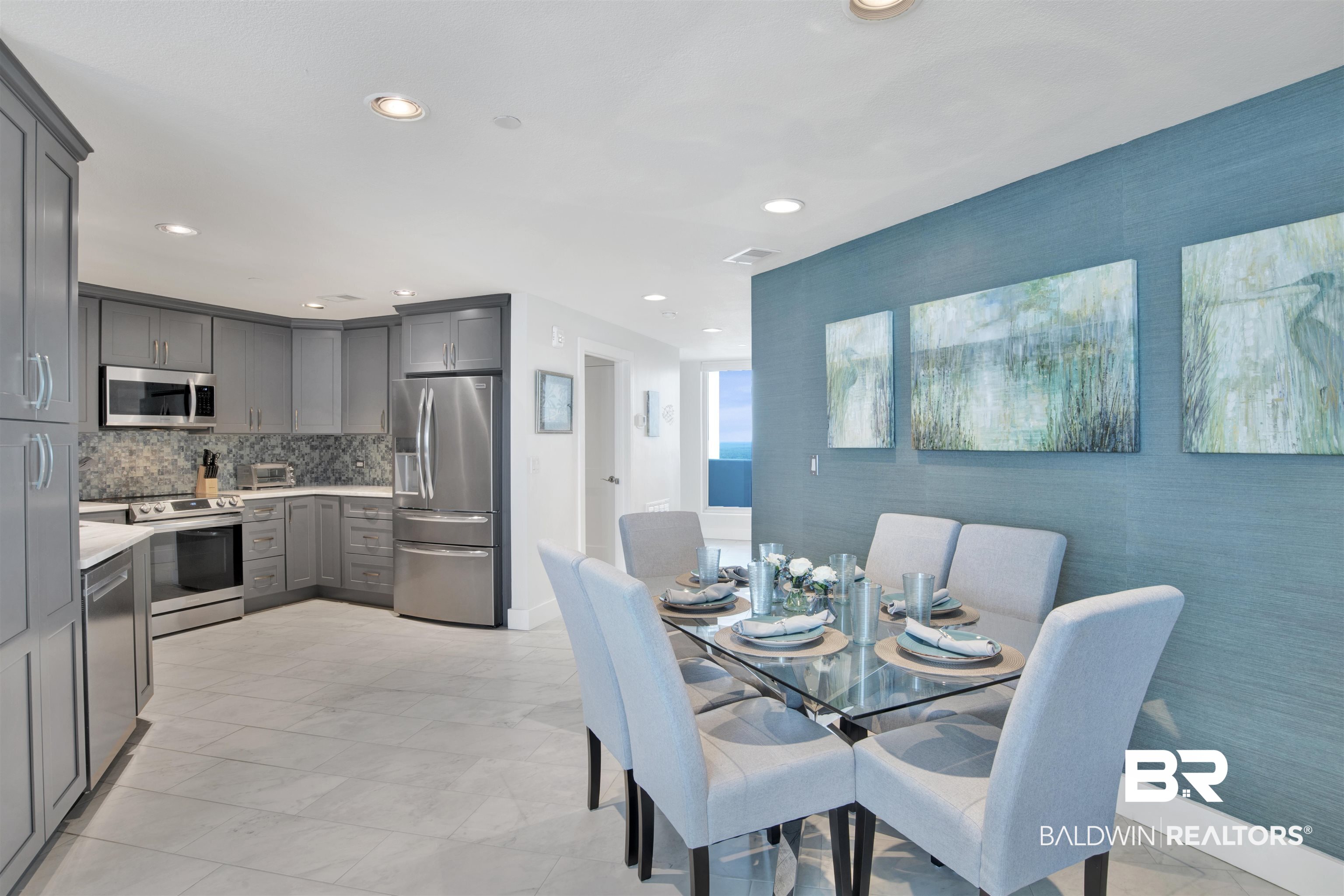 Open kitchen and dining area with gray cabinetry, a set glass table, and a glimpse of the ocean.