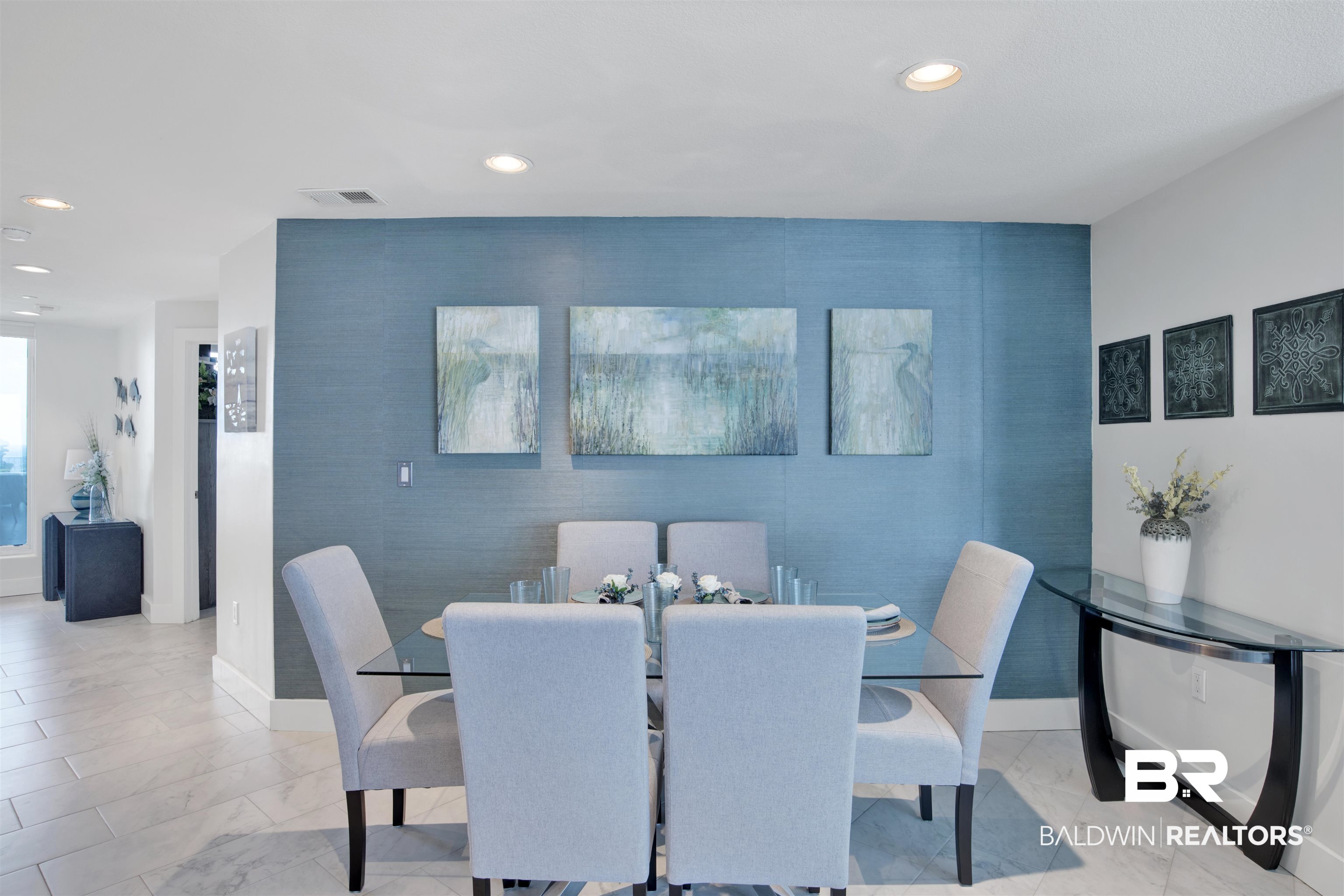 Dining room with a glass table, pale upholstered chairs, and blue accent wall with abstract artwork.