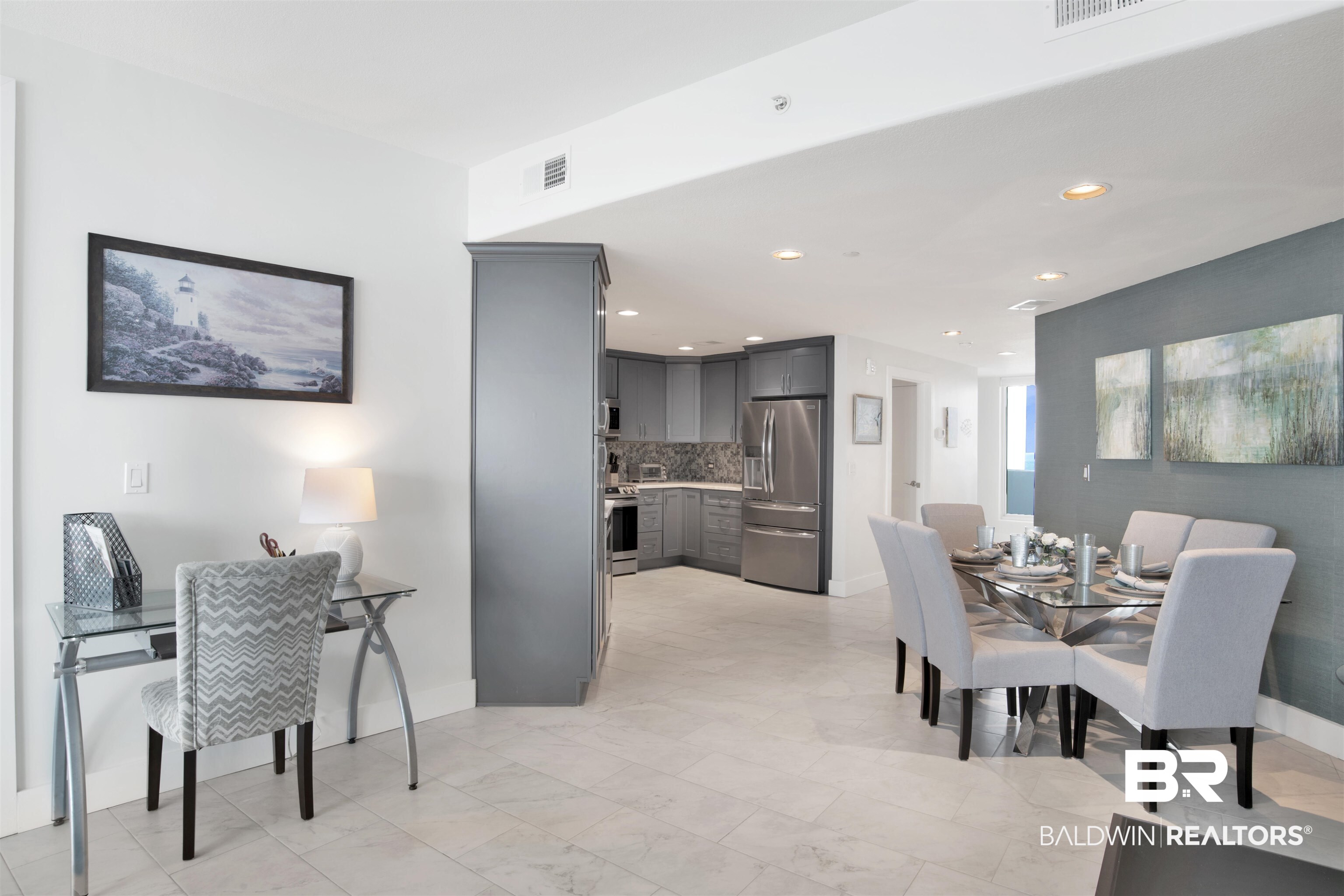 Open condo dining area and kitchen with gray cabinets, stainless steel appliances, and a glass dining table.