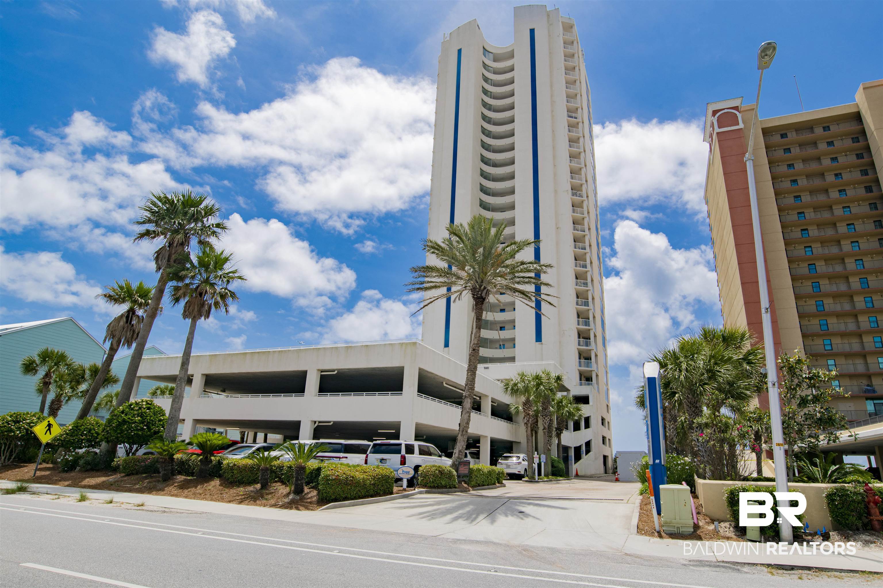 White condominium tower with curved balconies, blue accents, a parking garage, and palm trees.