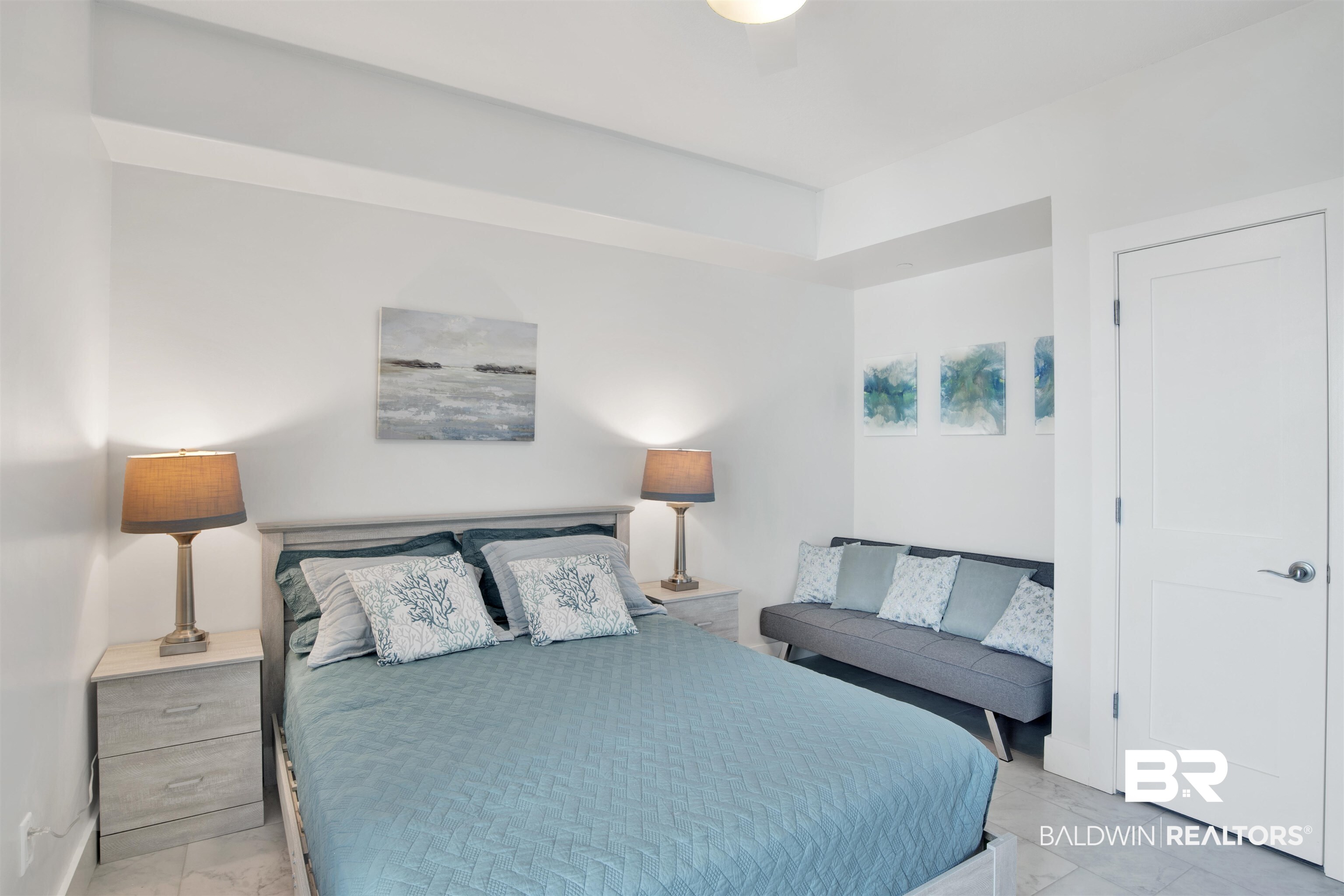 Bedroom with a teal quilted bed, matching lamps, coastal artwork, and a gray sofa.