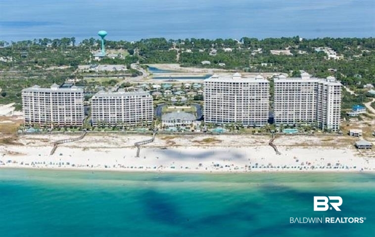 Aerial view of beachfront condominium towers beside a wide sandy beach and turquoise water.