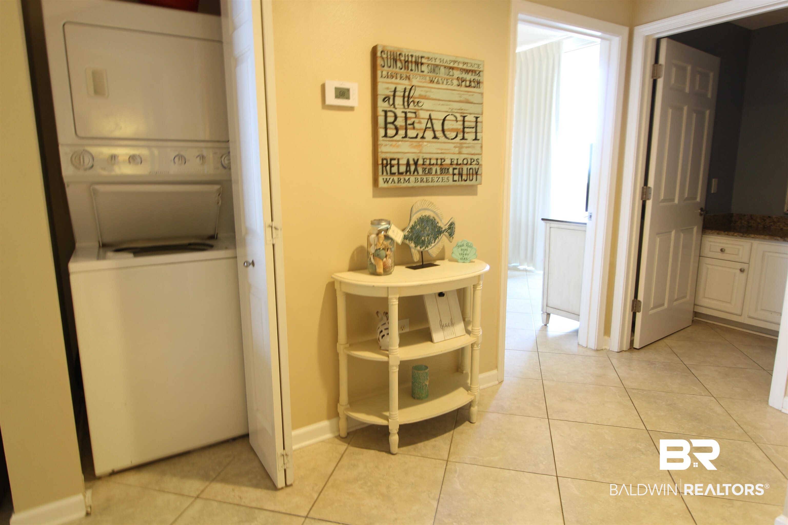 Laundry area with a stacked washer and dryer beside a beach-themed console table and tiled hallway.