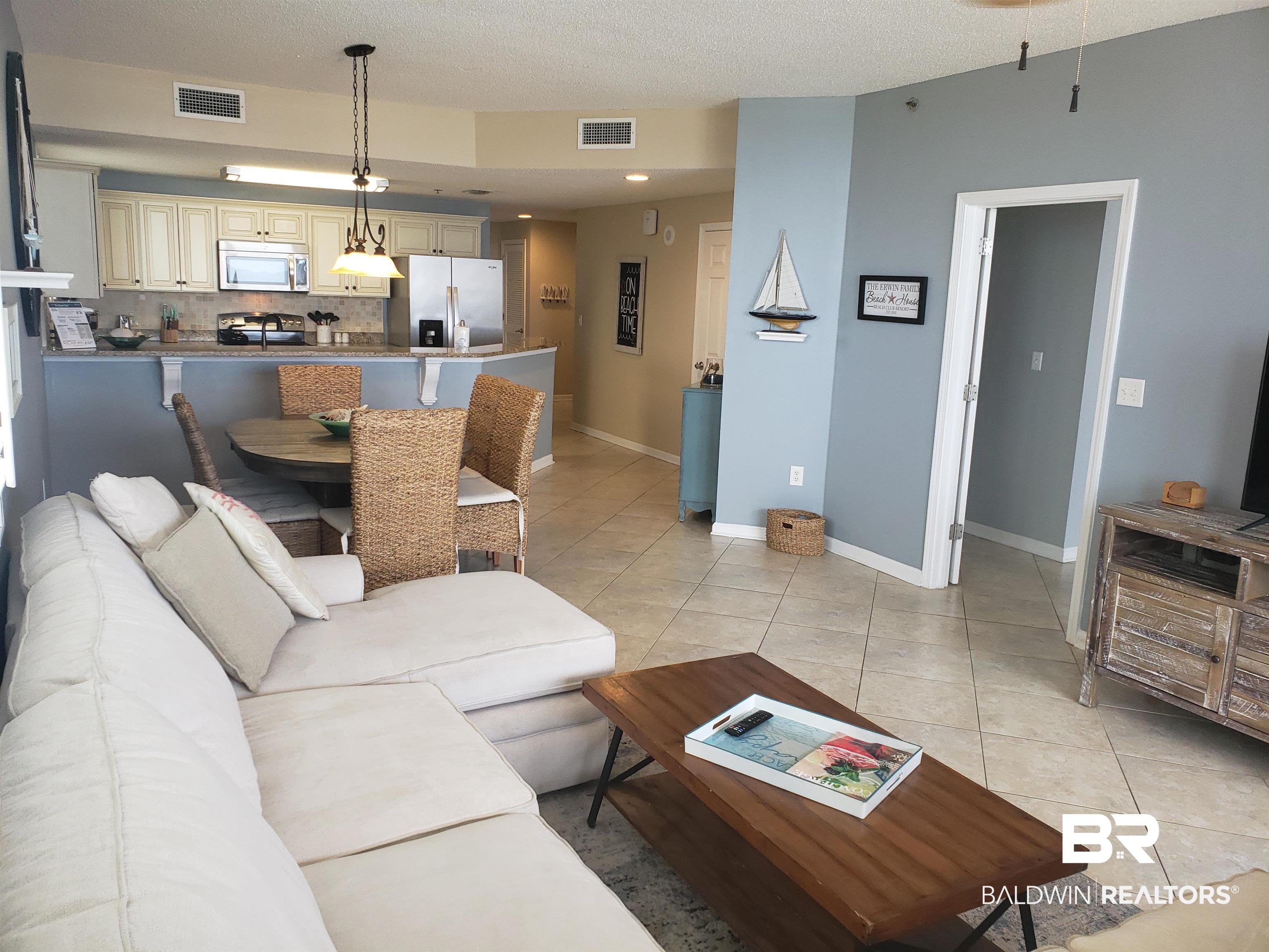 Open-plan living room with white sectional, wicker dining chairs, and kitchen breakfast bar.