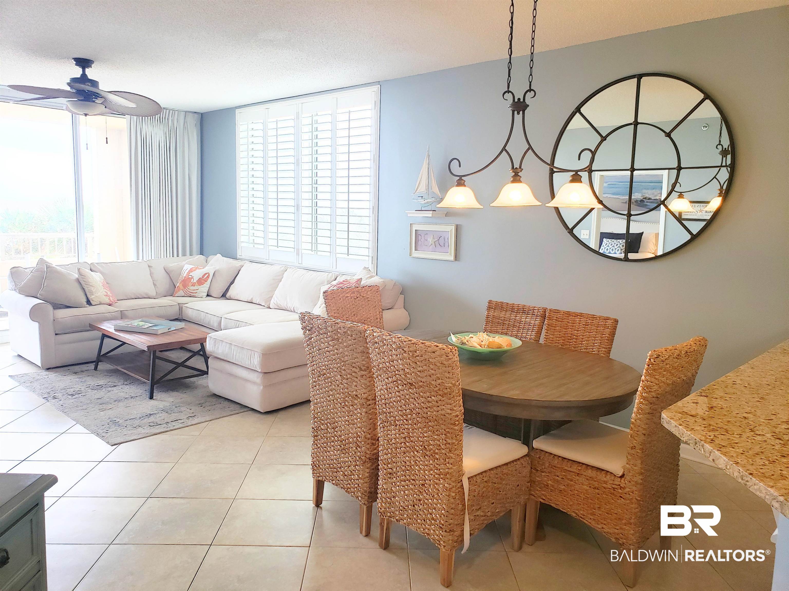 Furnished living and dining area with a white sectional, woven chairs, and a large round mirror.