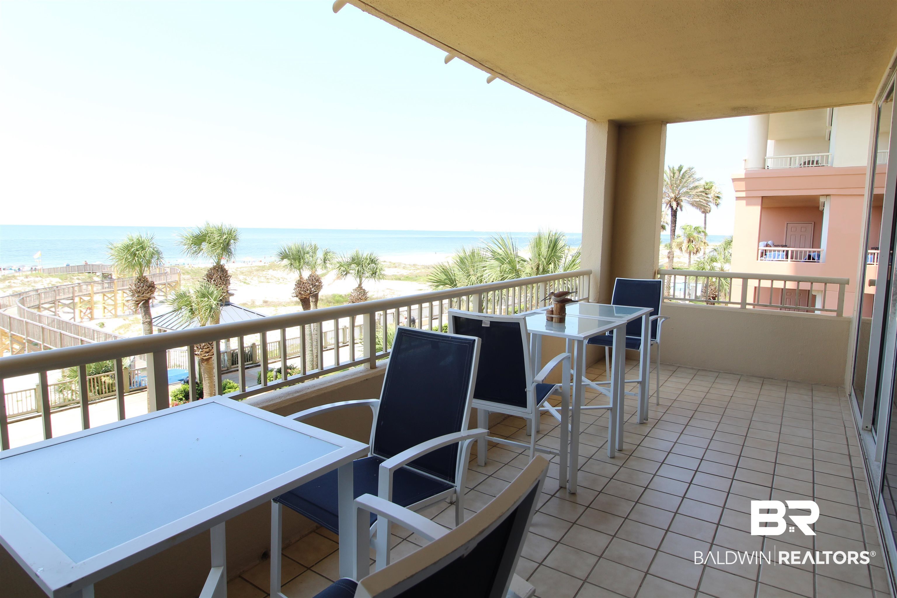 Furnished balcony overlooking the Gulf beach, with palm trees and a neighboring condominium building.