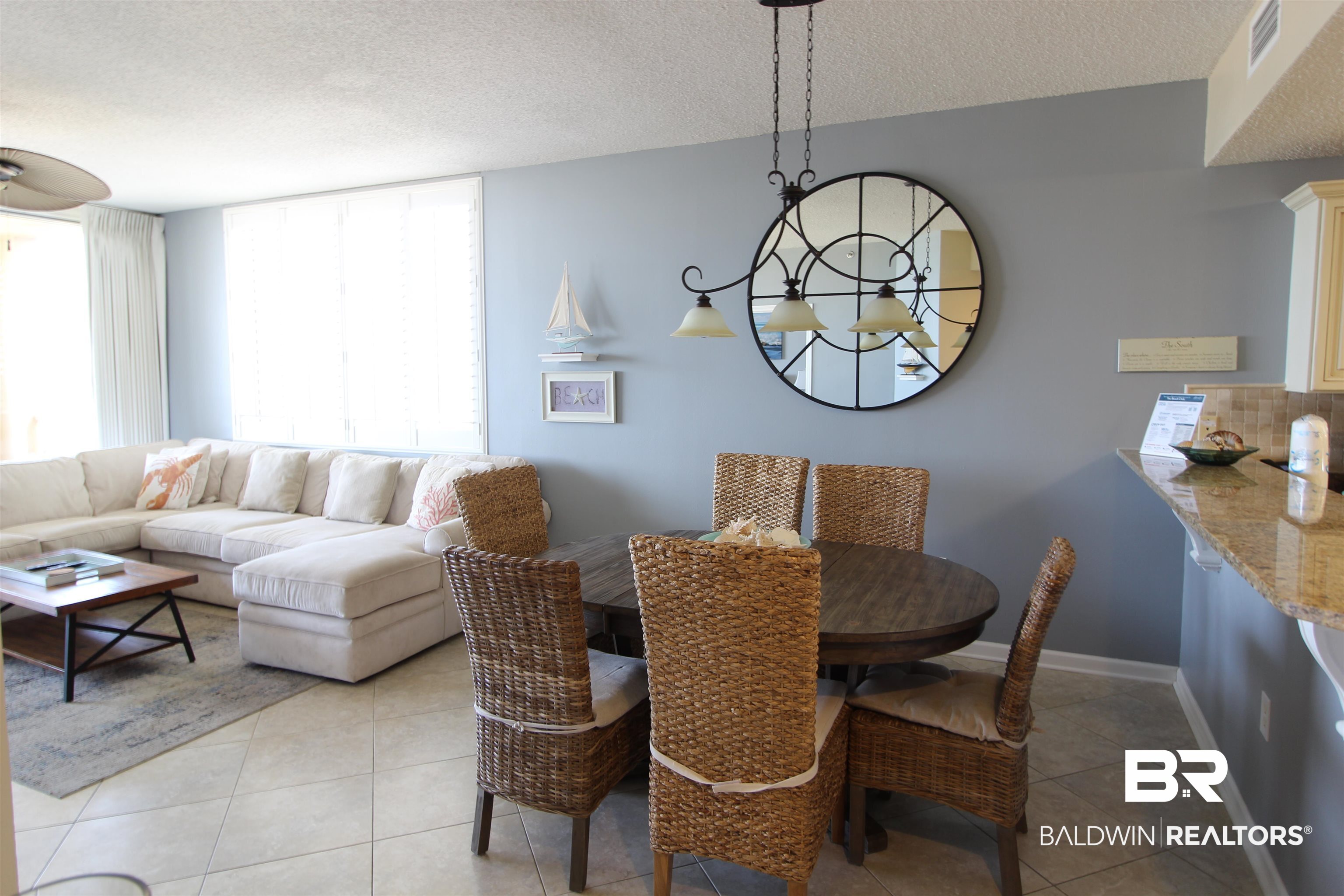 Living and dining area with a white sectional, wicker chairs, and a large round wall mirror.