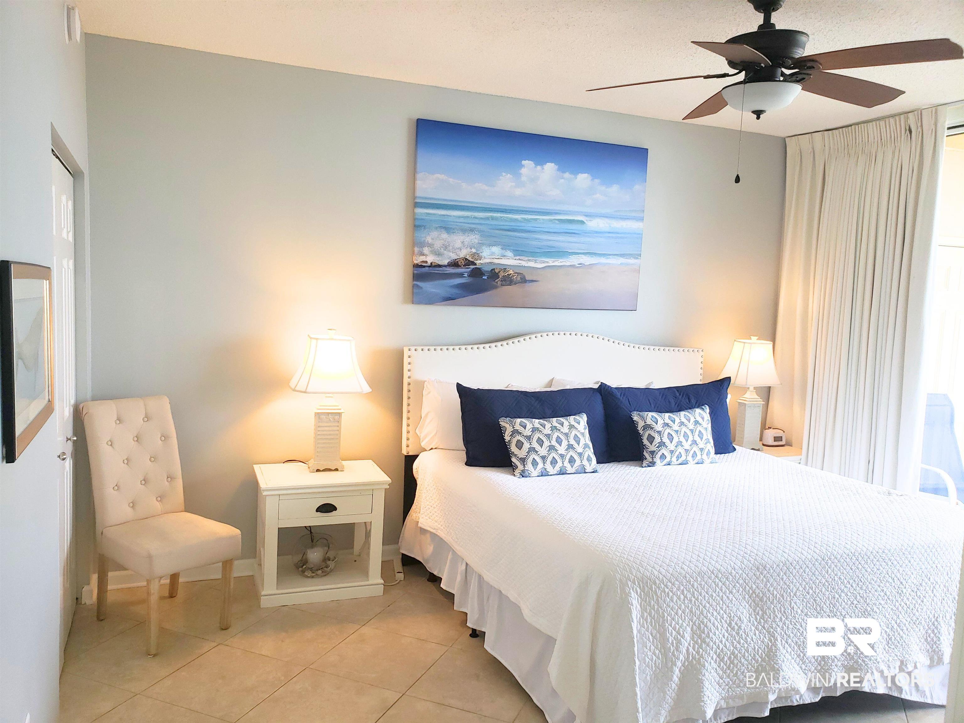 Furnished bedroom with a white bed, navy pillows, beach artwork, and matching bedside lamps.