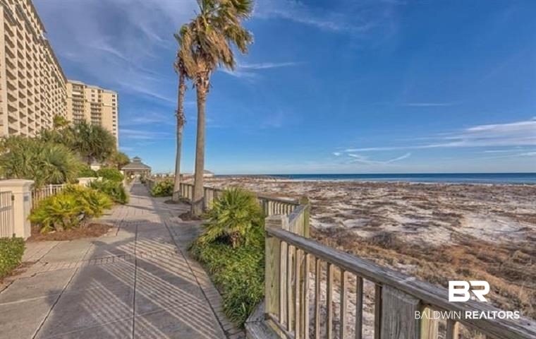 Beachfront boardwalk beside coastal condos, with palm trees and a wide Gulf shoreline.