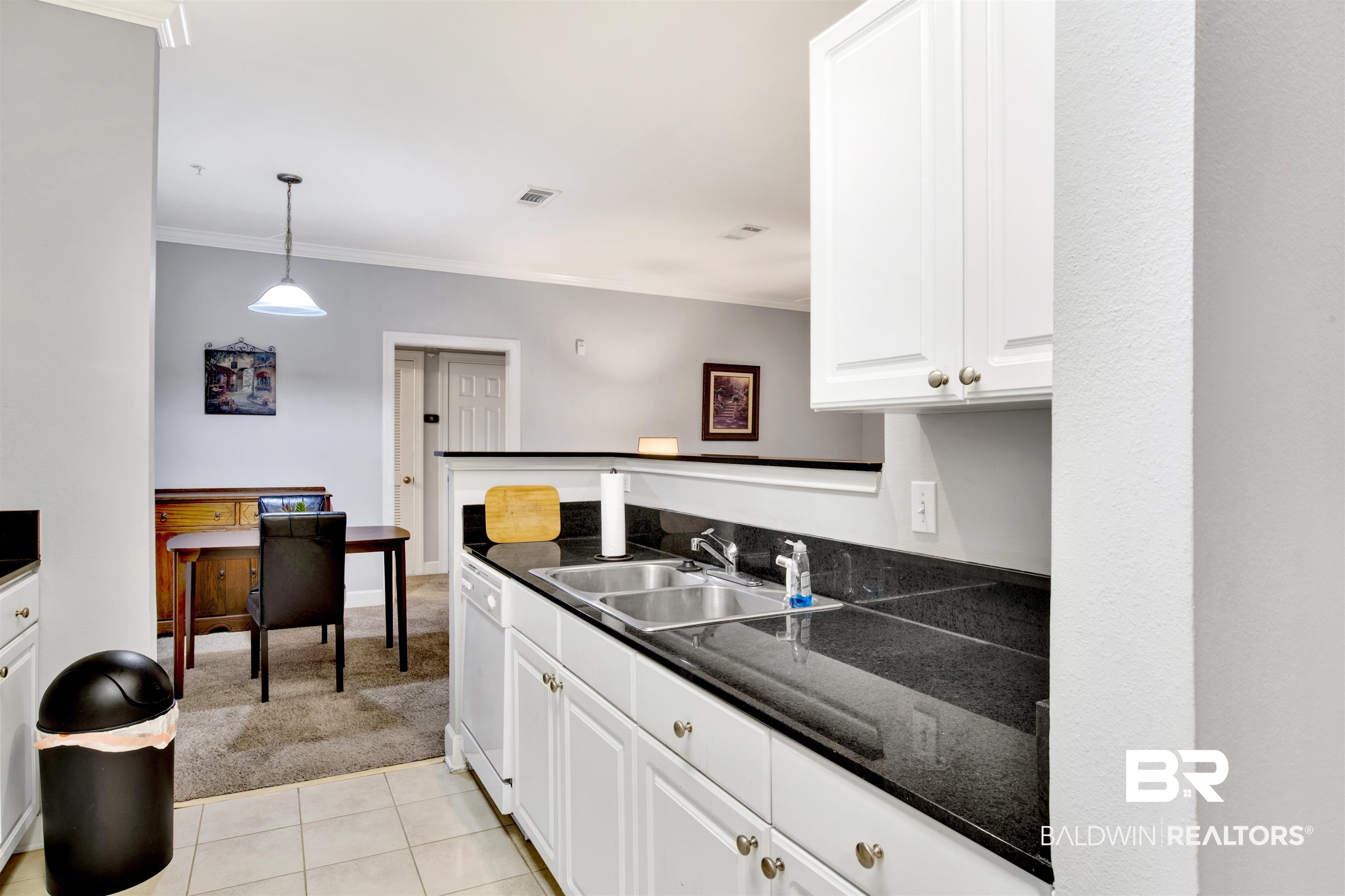 Kitchen with white cabinets, dark countertops, double sink, and a dining table beyond.