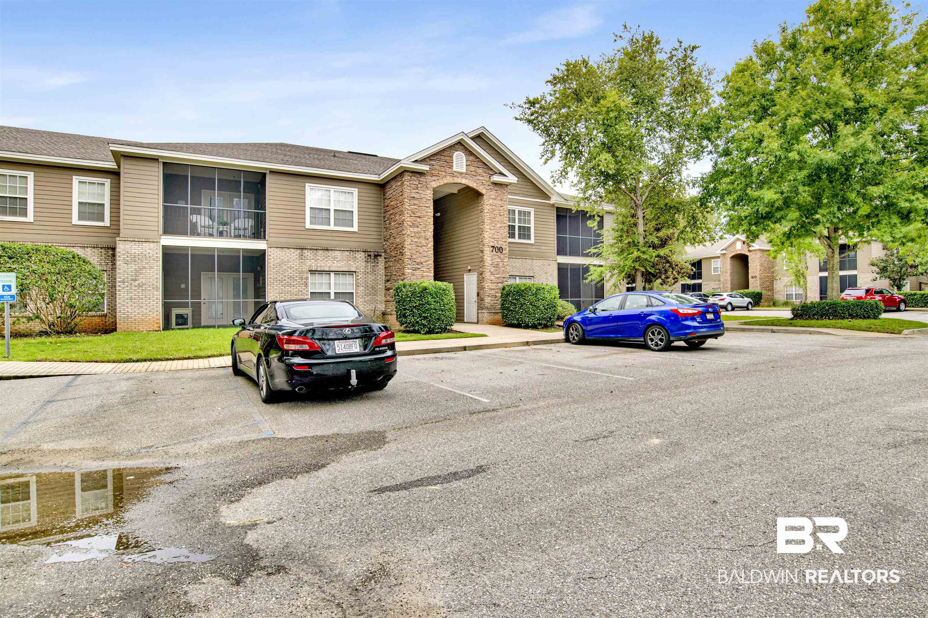 Two-story apartment building with a brick arched entrance, screened balconies, and cars in the parking lot.