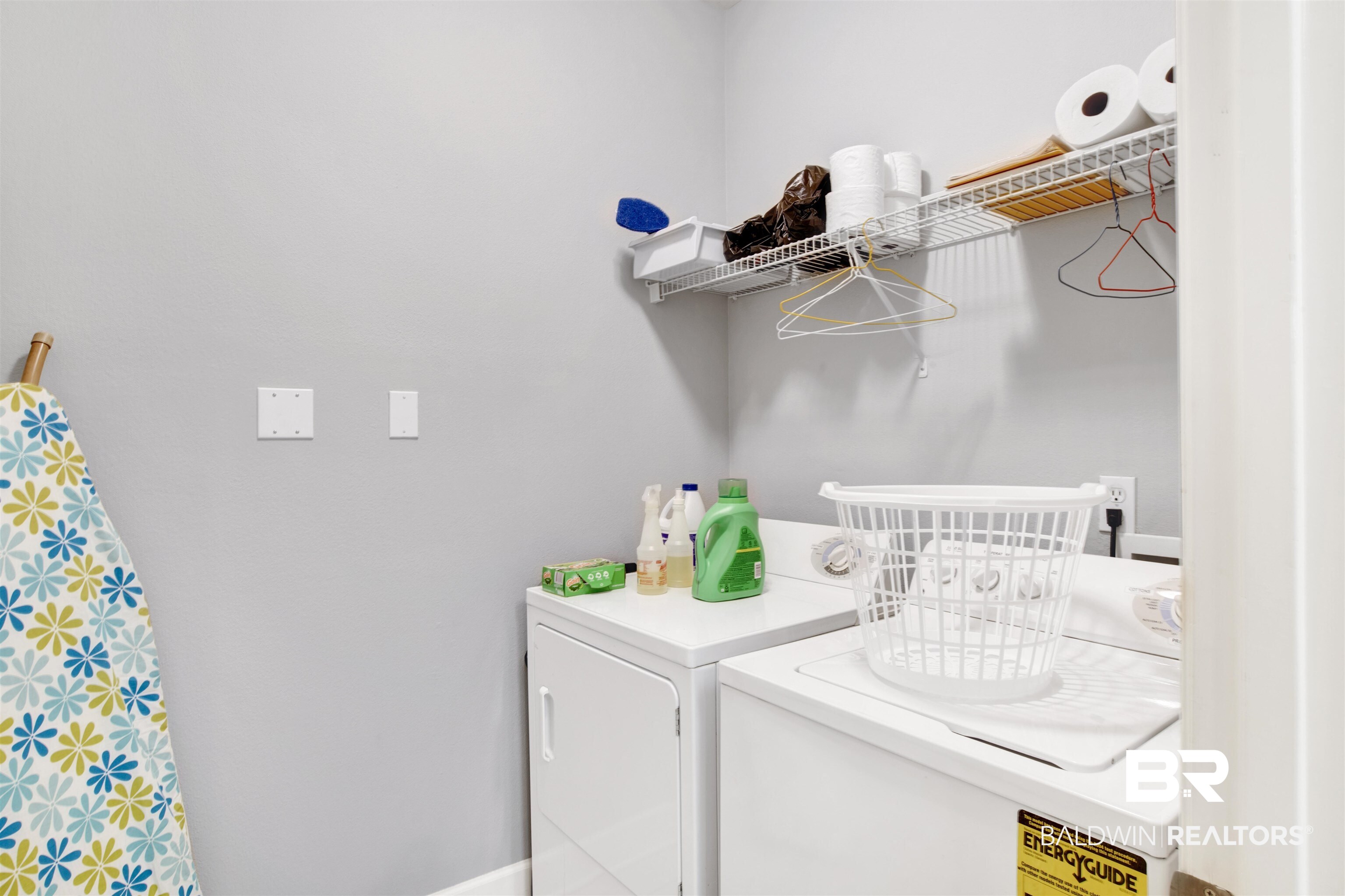 Laundry room with white washer and dryer, a wire shelf, and a floral ironing board.