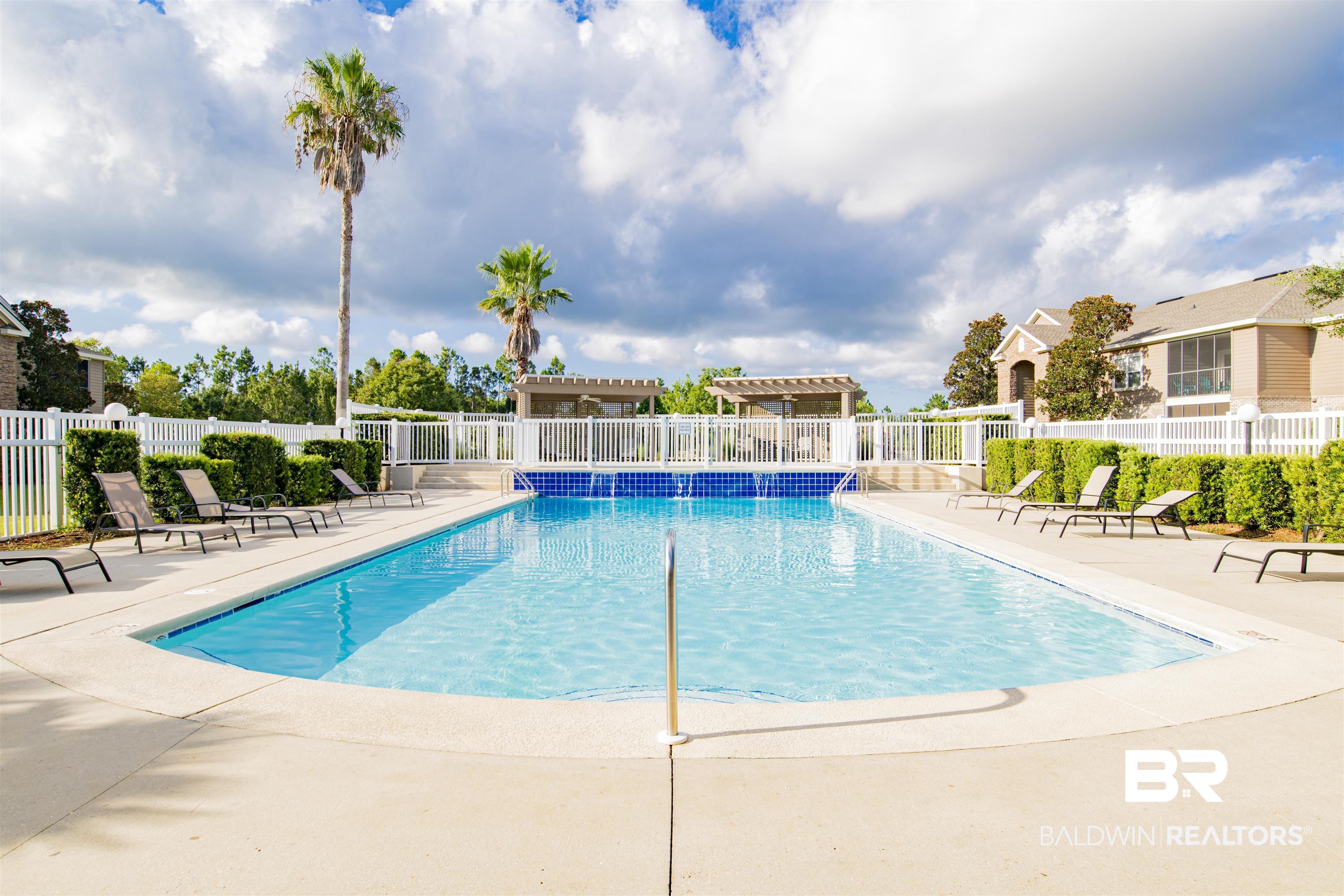 Outdoor swimming pool with lounge chairs, palm trees, and a white fence.