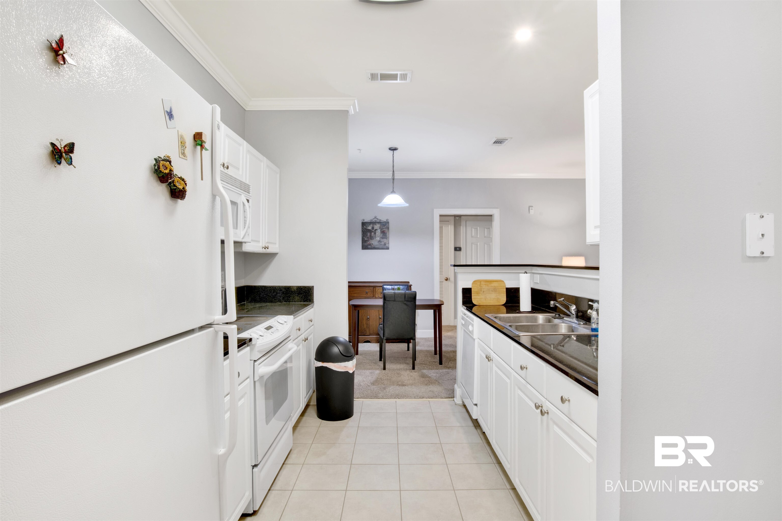 Galley kitchen with white cabinetry, dark countertops, and a dining area beyond.