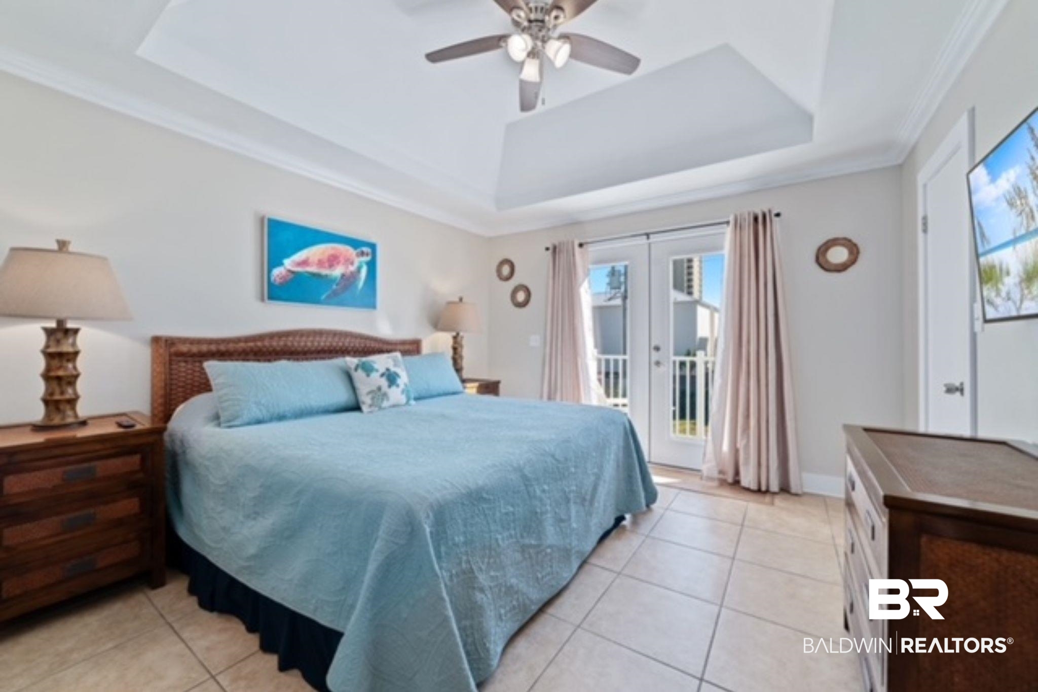 Bedroom with a king-size bed in pale blue bedding, sea turtle artwork, and glass doors to a balcony.