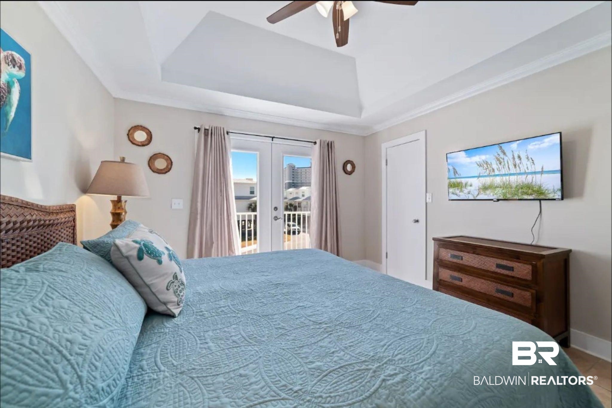 Bedroom with a pale blue quilted bed, woven furnishings, tray ceiling, and glass doors to a balcony.