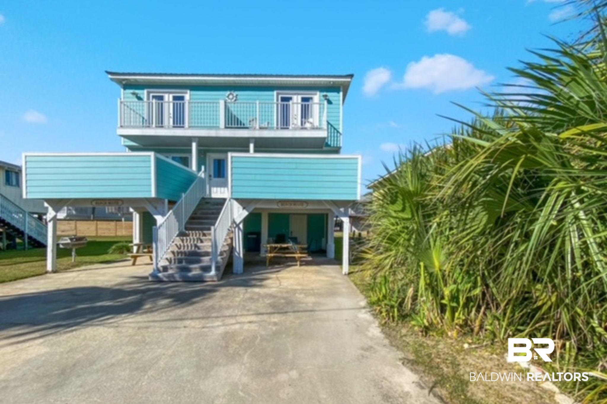 Raised turquoise beach house with a white-railed balcony, central staircase, and tropical grasses along the driveway.