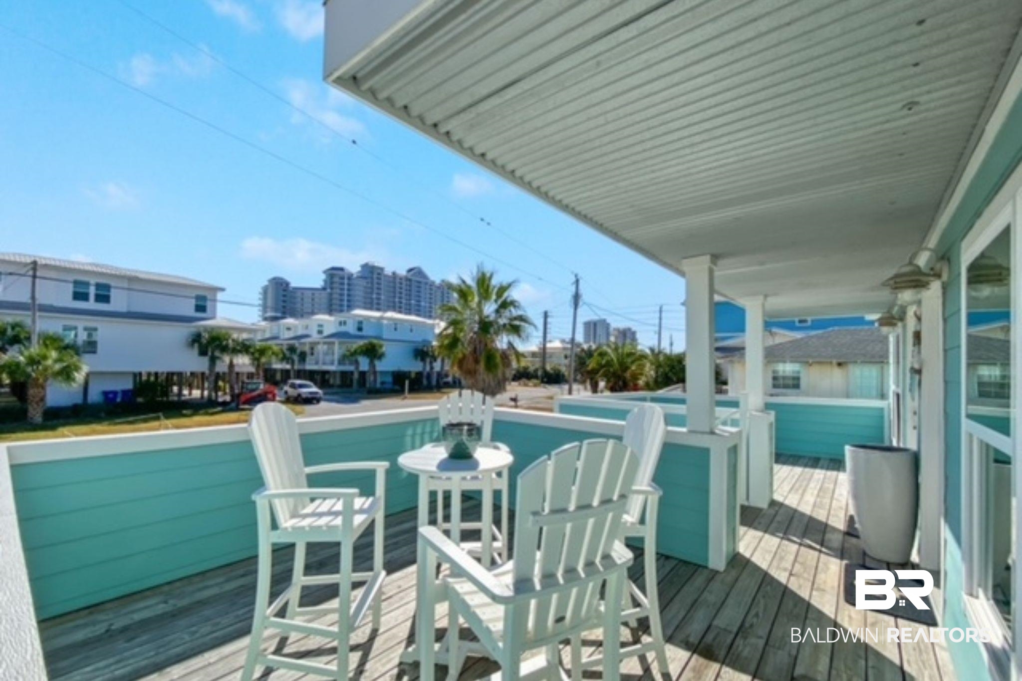 Covered deck with turquoise railings and white Adirondack chairs overlooking palm trees and nearby buildings.