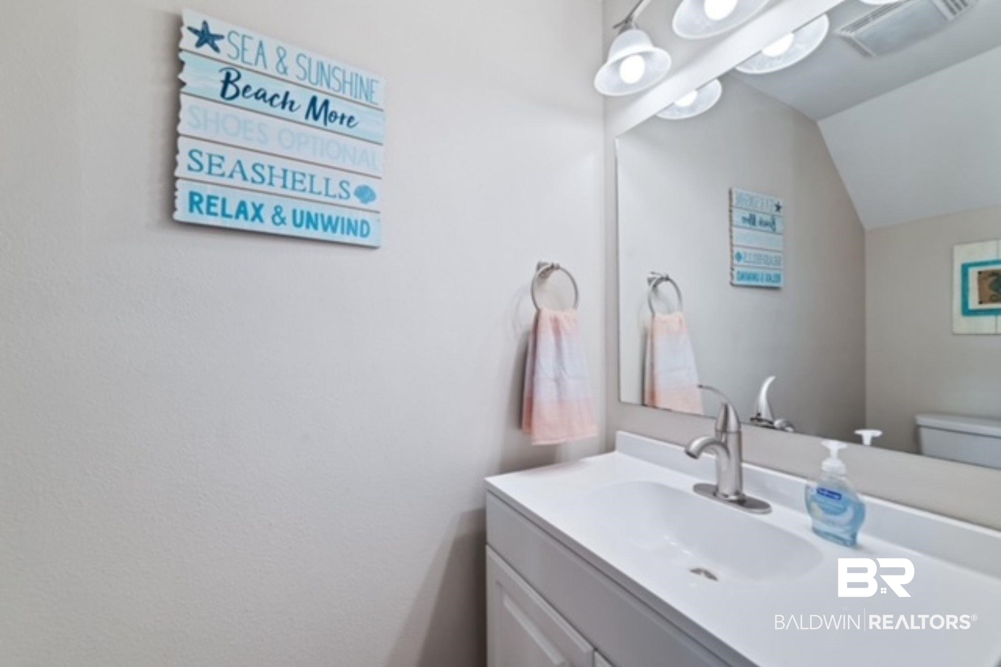 Bathroom vanity with a white sink, large mirror, pink towel, and coastal-themed wall sign.