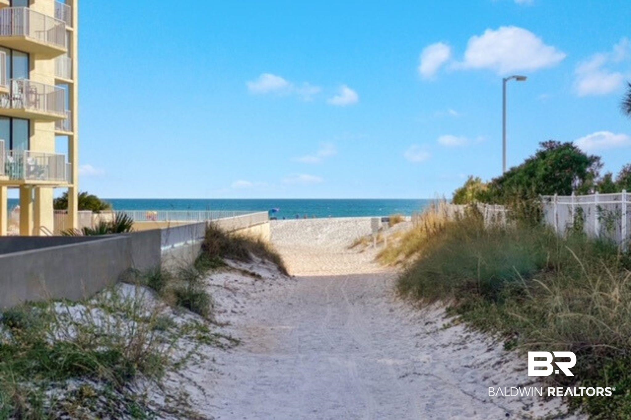 Sandy beach access path leads to the Gulf, bordered by dune grasses and a condominium building.
