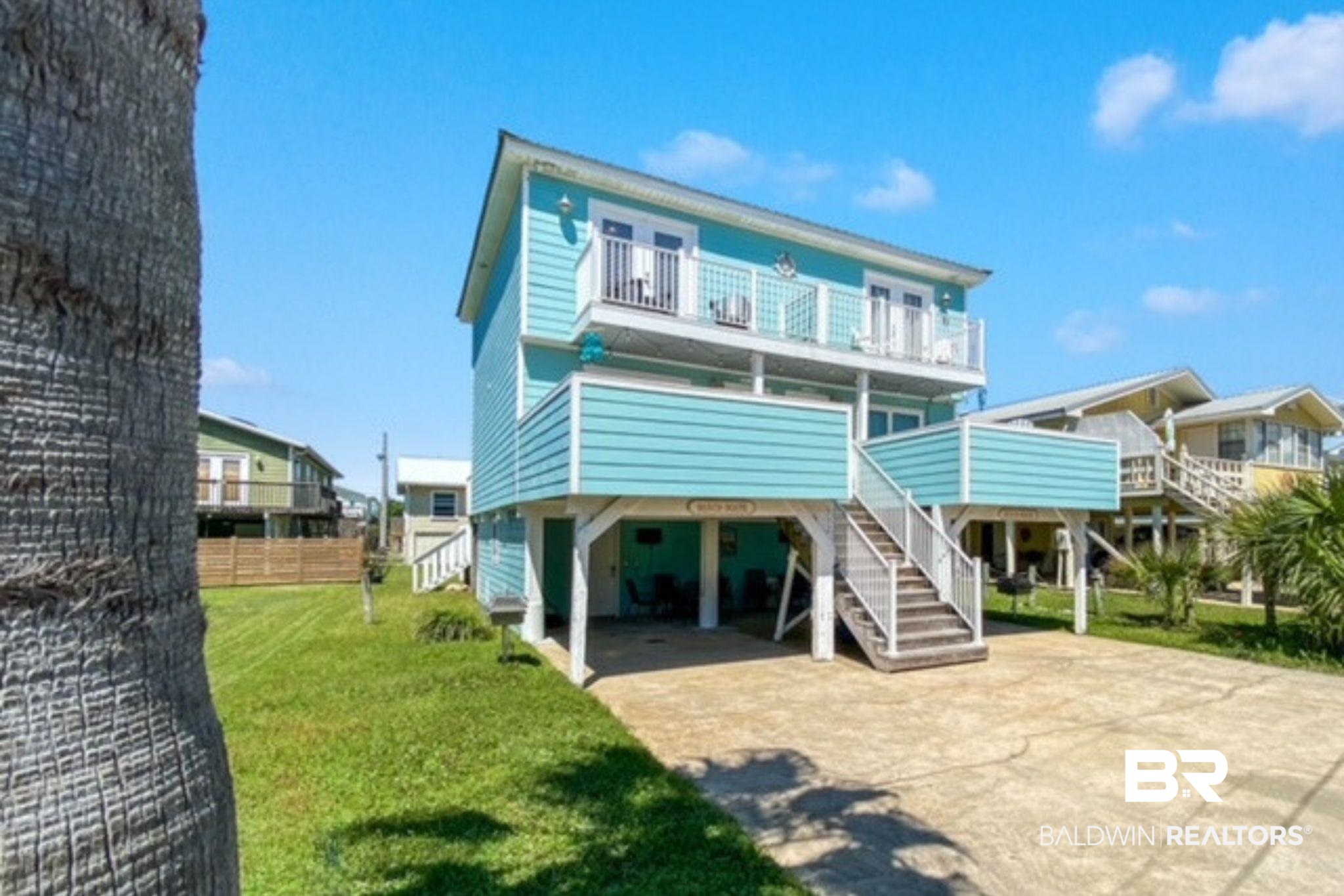 Raised turquoise beach house with white balconies and exterior stairs beside a paved driveway.