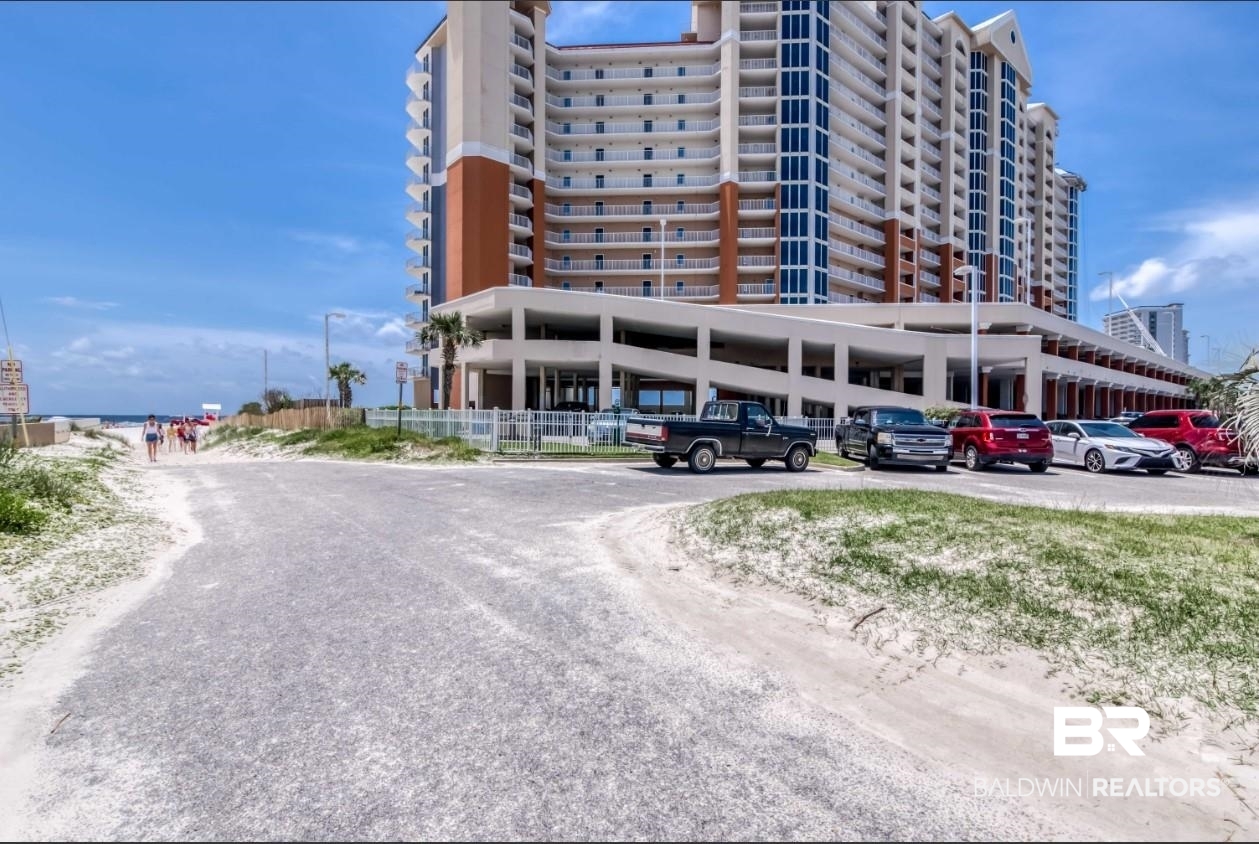 Multistory beachfront condominium with curved balconies, parking, and a sandy path leading toward the water.