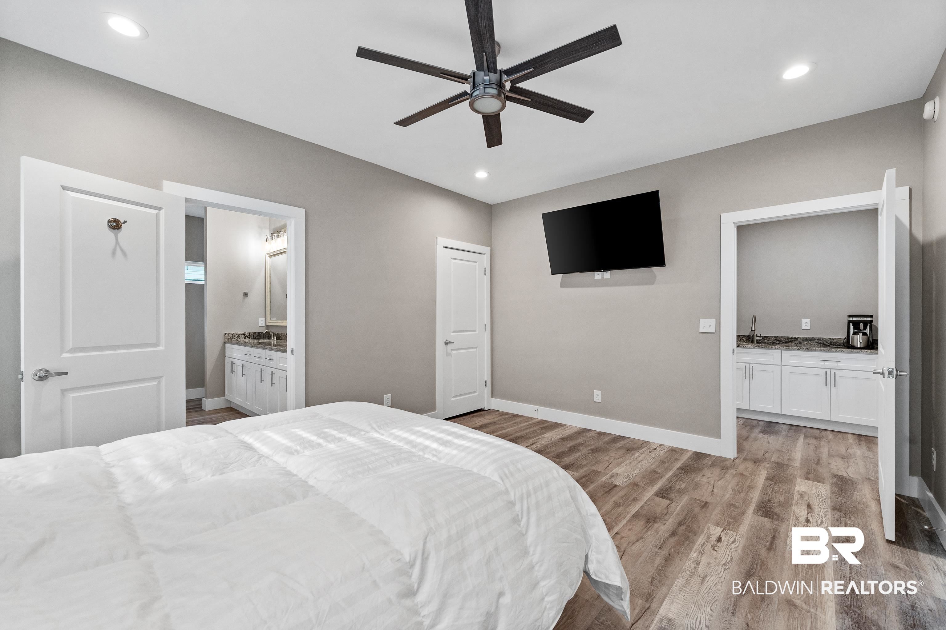 Bedroom with a white bed, dark ceiling fan, wall-mounted TV, and adjoining vanity areas.