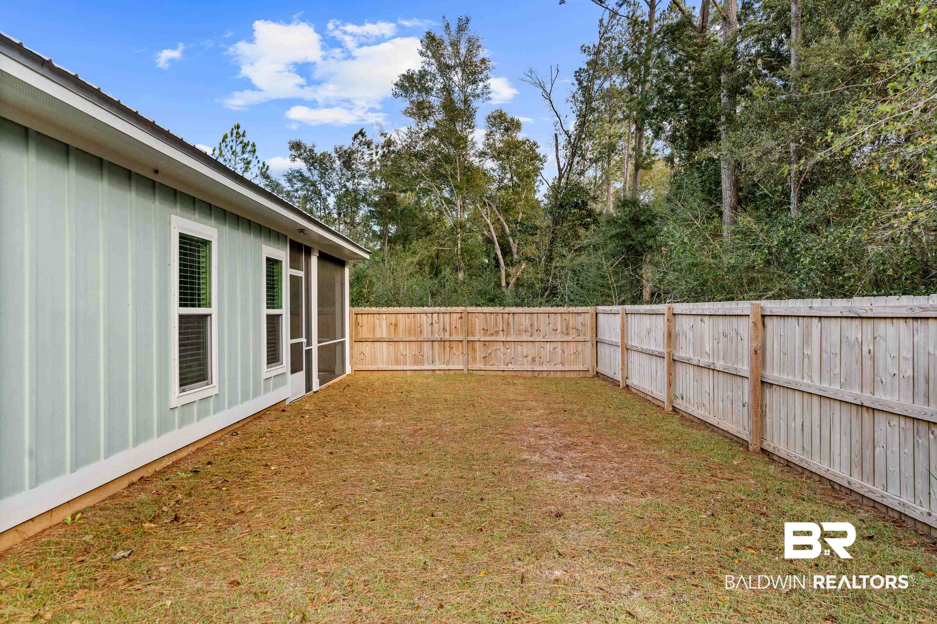 Fenced backyard beside a pale green home, with a screened porch and mature trees beyond the fence.