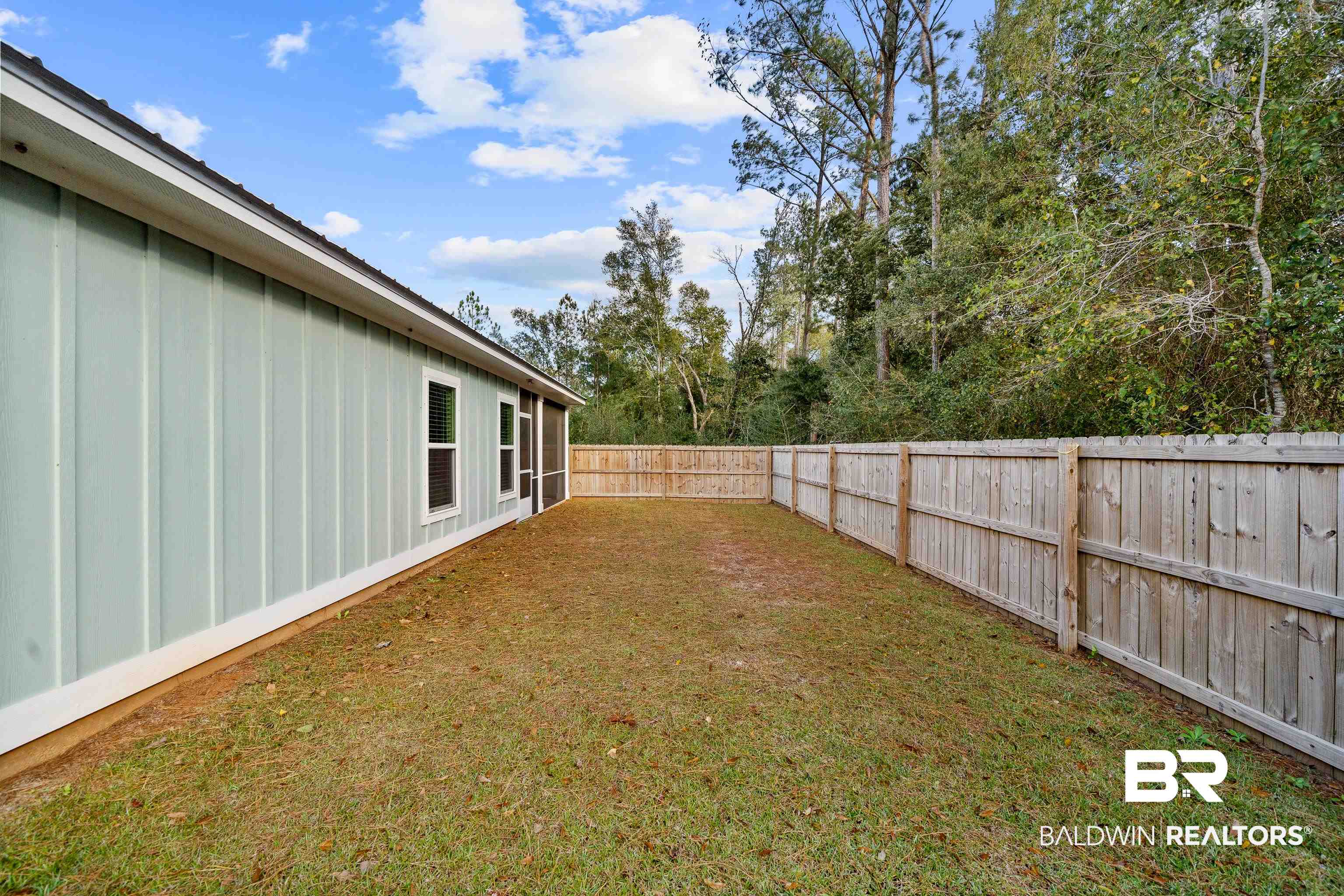 Fenced backyard beside a pale green-sided home, with a patchy lawn and mature trees beyond.