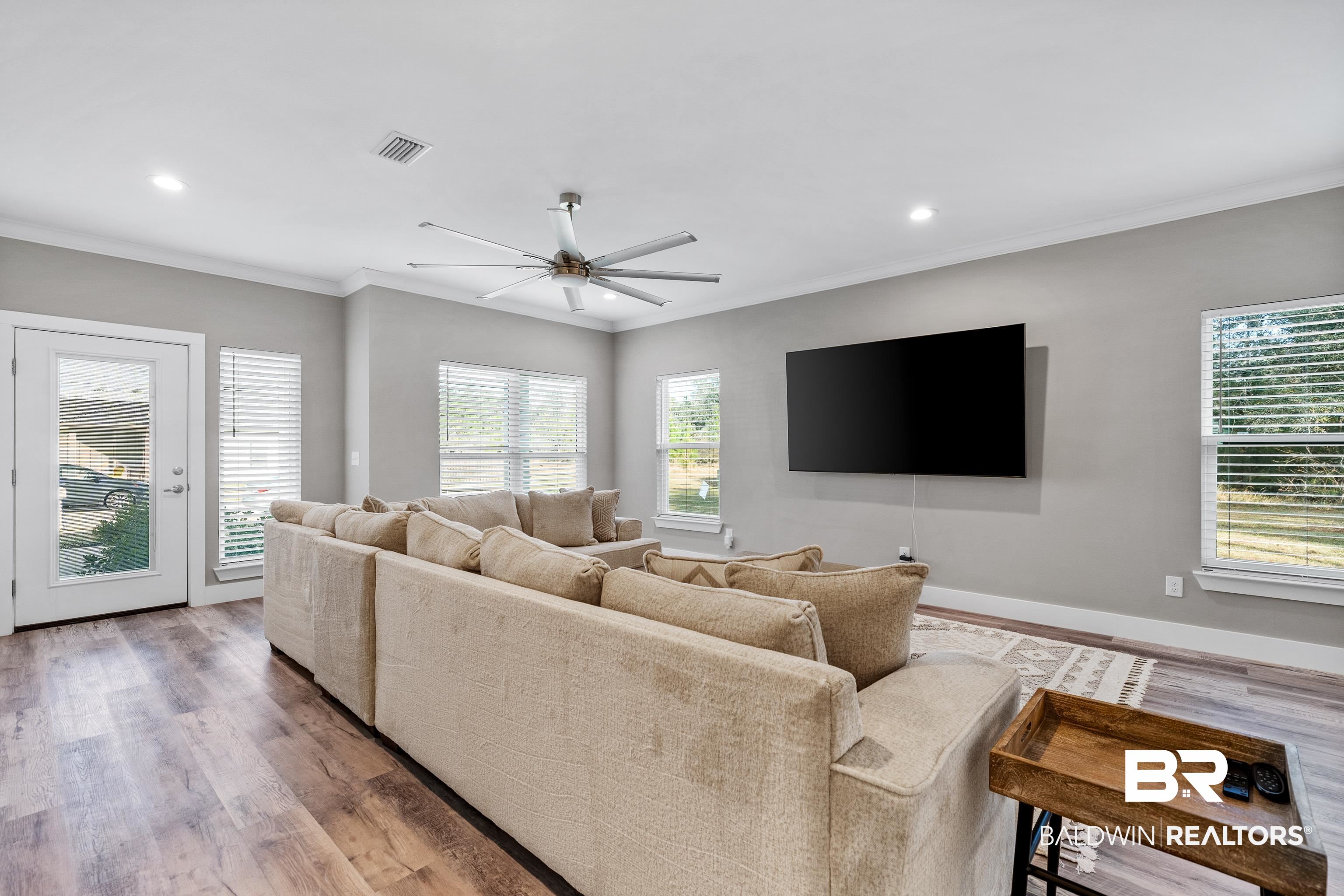 Living room with a beige sectional sofa, wall-mounted television, and large windows with blinds.