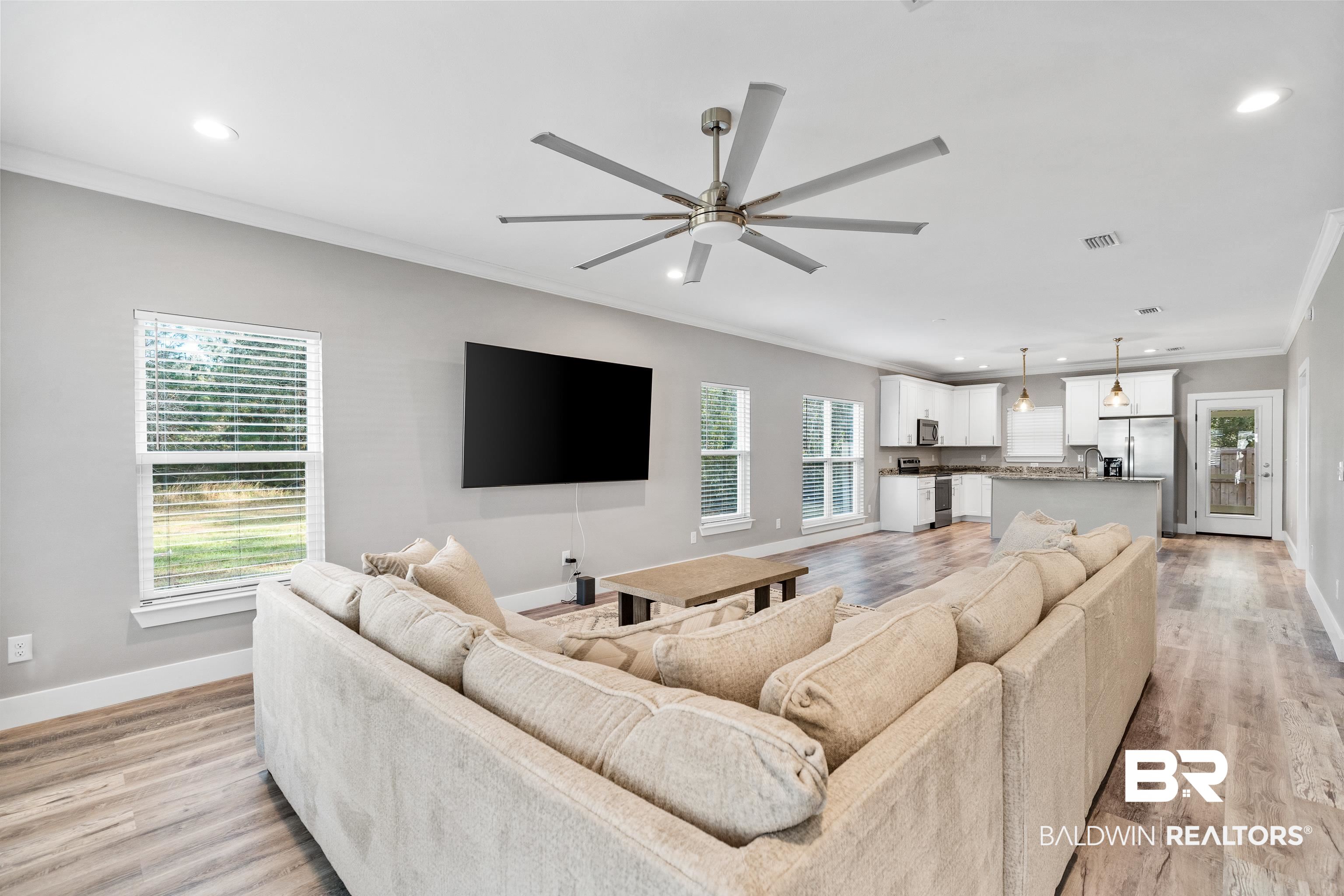 Open-plan living room and kitchen with a large ceiling fan, sectional sofa, and wood-look flooring.