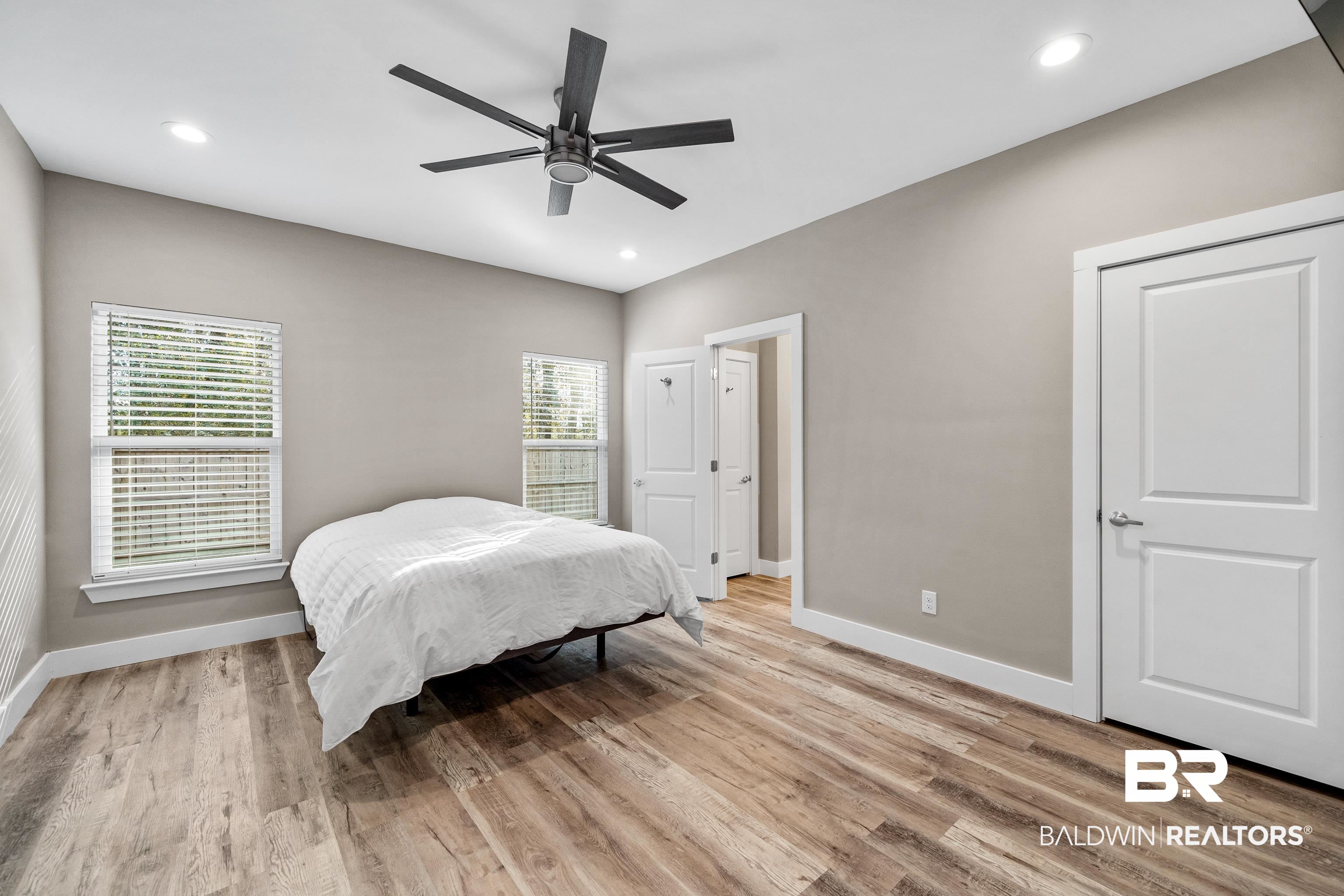 Bedroom with white bedding, two windows with blinds, wood-look flooring, and a dark ceiling fan.