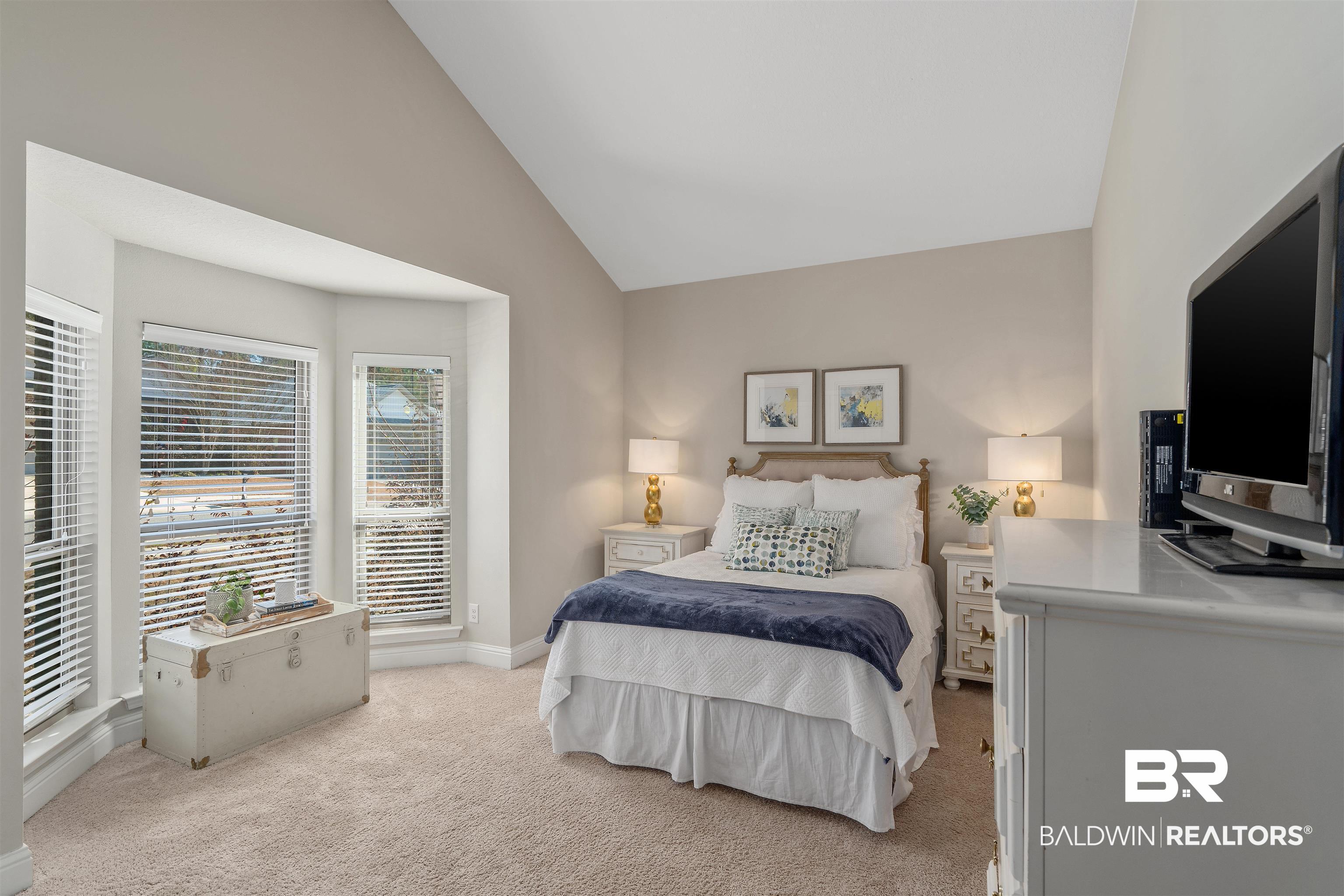 Furnished bedroom with a white bed, navy blanket, and bright bay windows with white blinds.