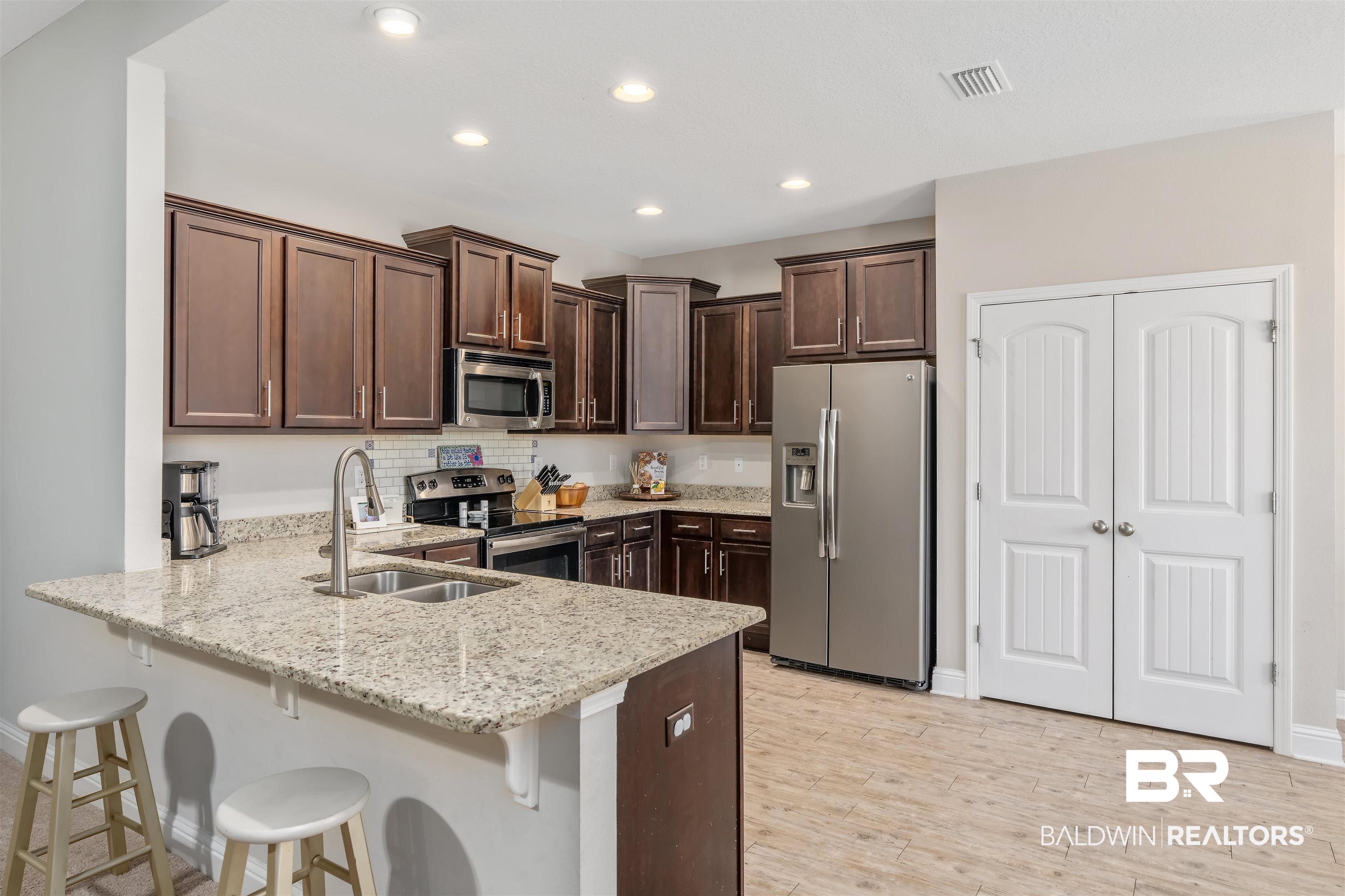 Kitchen with dark wood cabinets, granite breakfast bar, white stools, and stainless steel appliances.